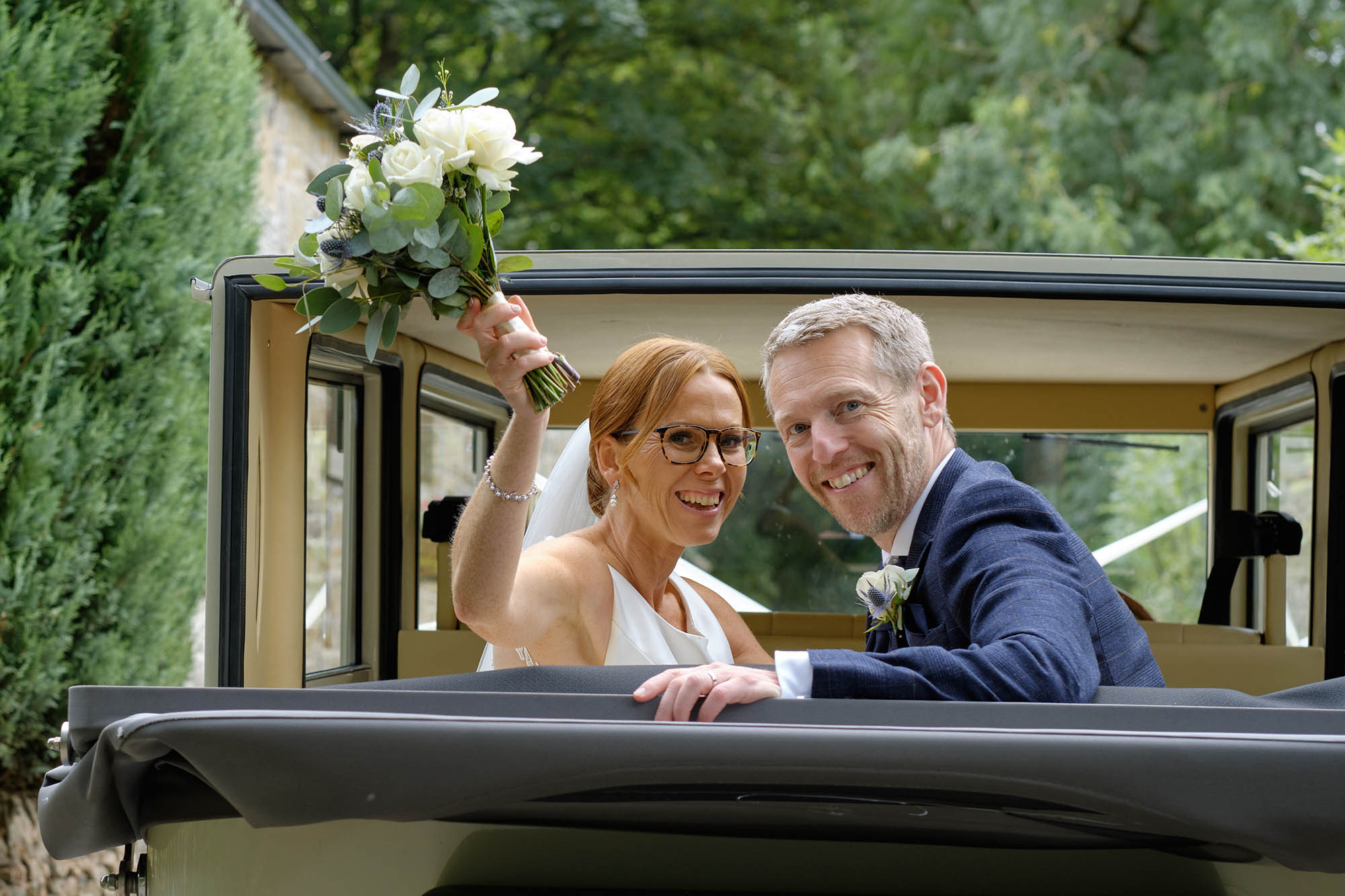 Bride and groom wave flowers and smile from the back of their vintage wedding car - Sheffield wedding photographer John Mottershaw