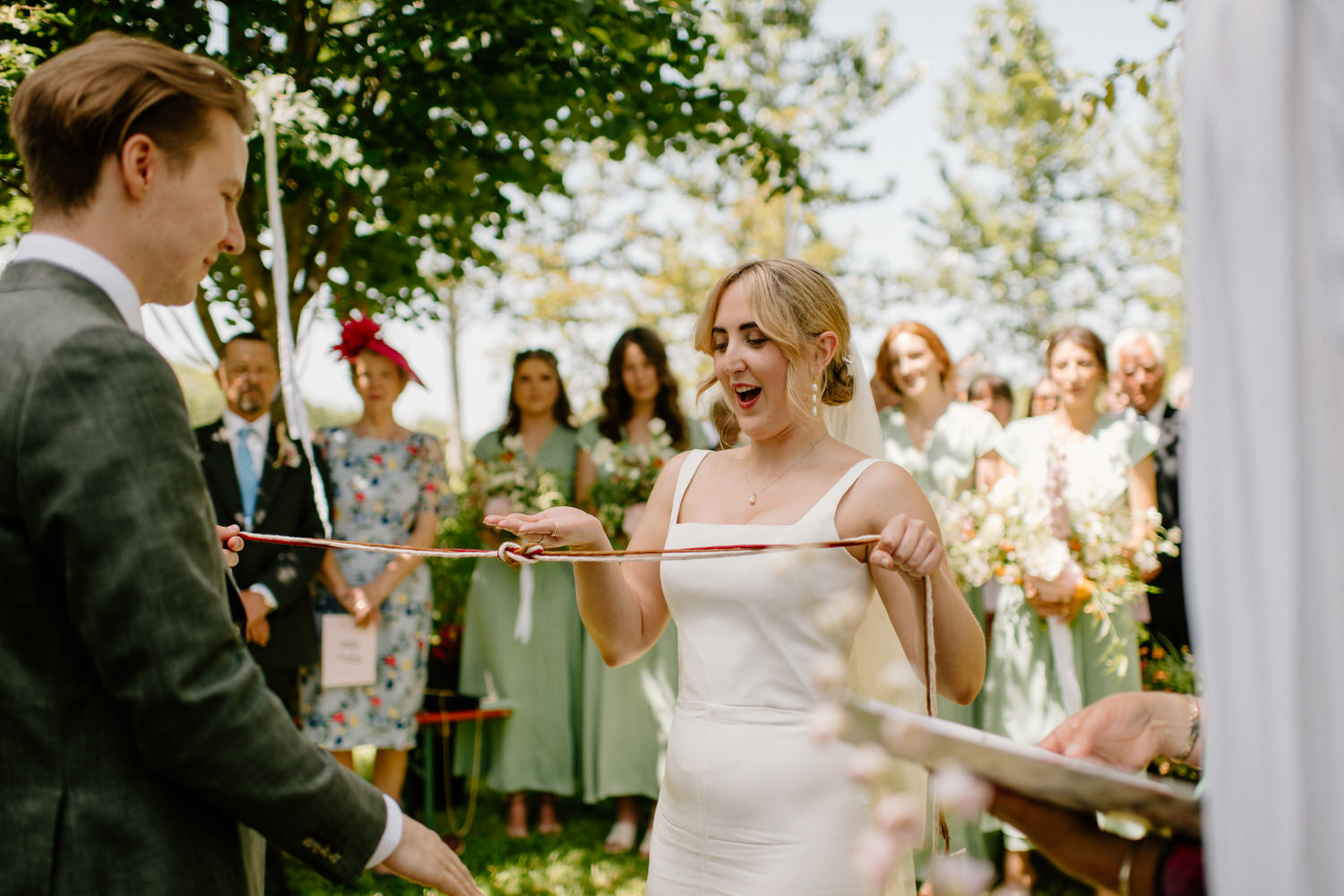Handfasting style wedding ceremony in the sunshine. The bride wears a simple modern halter neck dress. With Ellie Gillard Photography