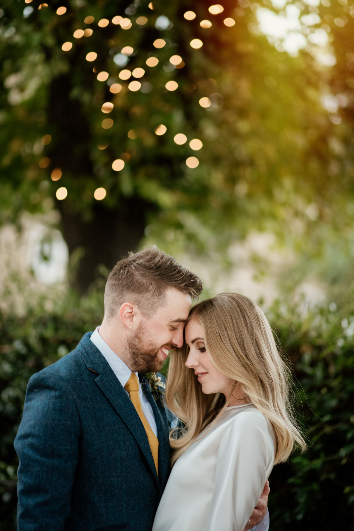 Romantic photo of a couple holding each other close on their wedding day. He's smiling; she looks blissful and content. There are fairy lights and trees in the background. By London wedding photographer Ellie Gillard