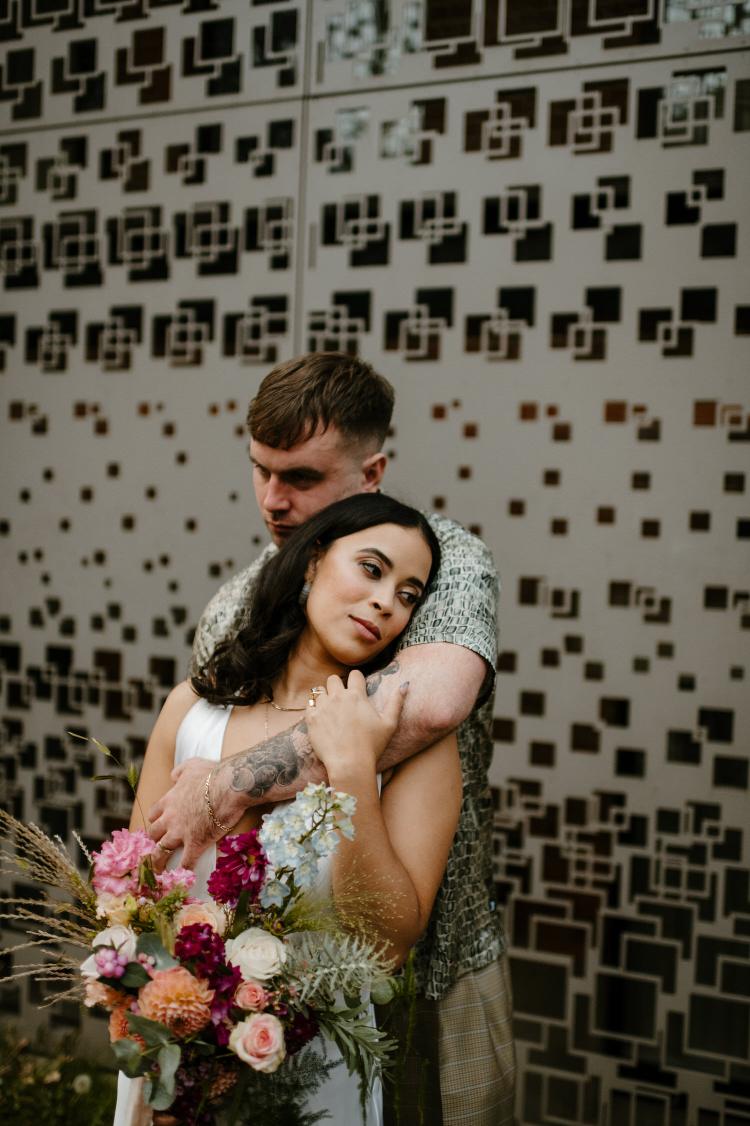 Modern alternative wedding photography by Ellie Gillard in London. A casually dressed groom holds his bride as she rests her head on his shoulder. She's carrying a bold red and pink bouquet