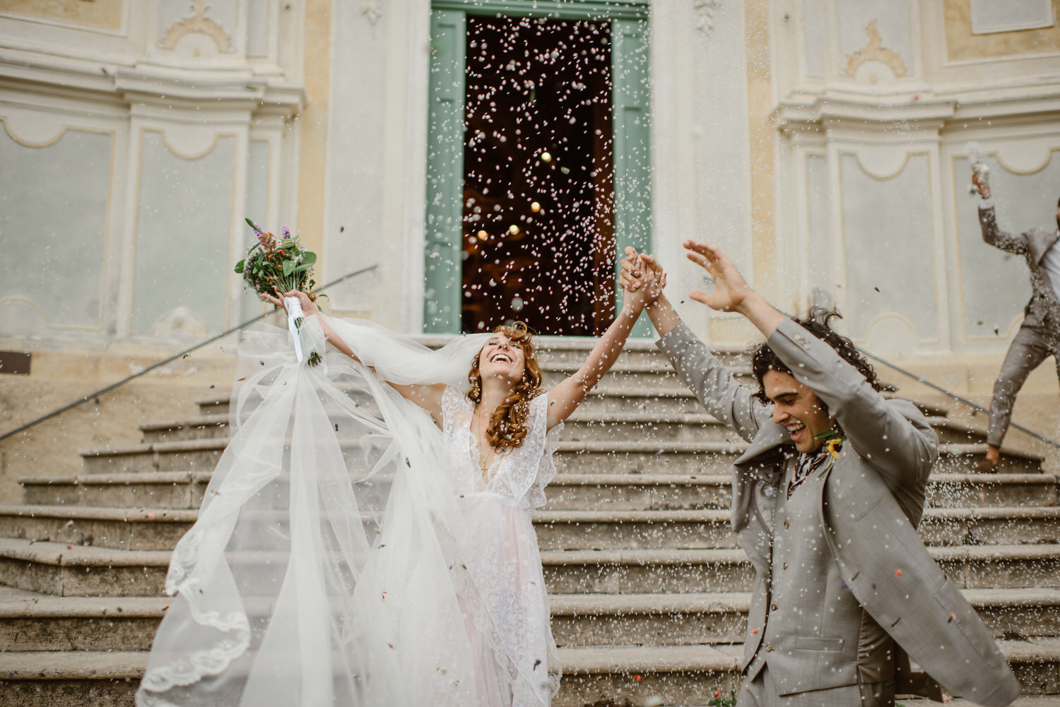 A bride and groom celebrate with their arms raised as a cloud of white confetti falls around them. She's in a white dress with a green bouquet and he's in a pale grey suit. They're on the steps on the ceremony venue. With Ellie Gillard Photography