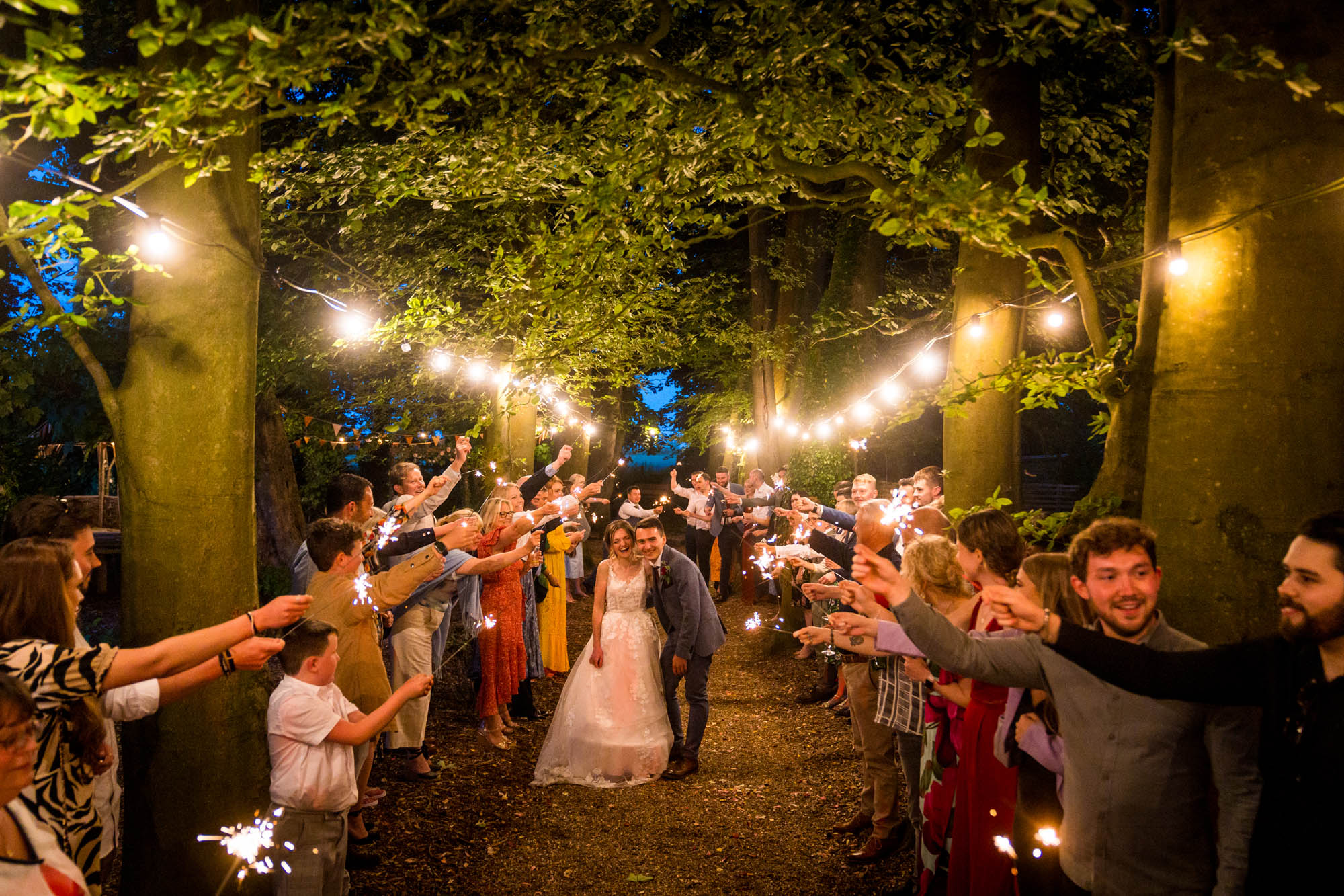 Newlyweds walking through their guests lined up with sparklers, in a woodland with festoon lighting