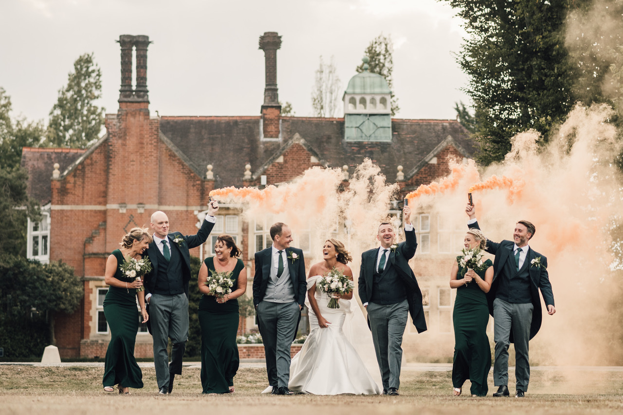 The bridal party at a wedding raise apricot coloured smoke bombs as they walk away from the wedding venue