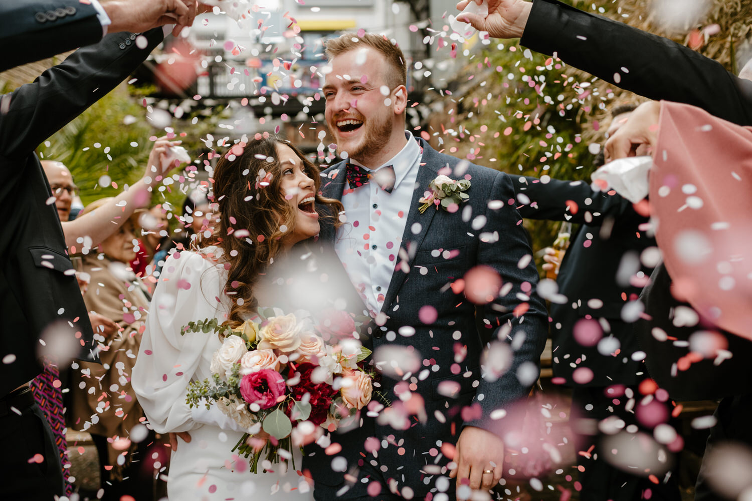 Joyful and colourful confetti shot of a bride and groom. She's wearing a long sleeved, minimal and vintage style white dress and he's in a blue suit and white shirt with a bowtie. By London wedding photographer Ellie Gillard
