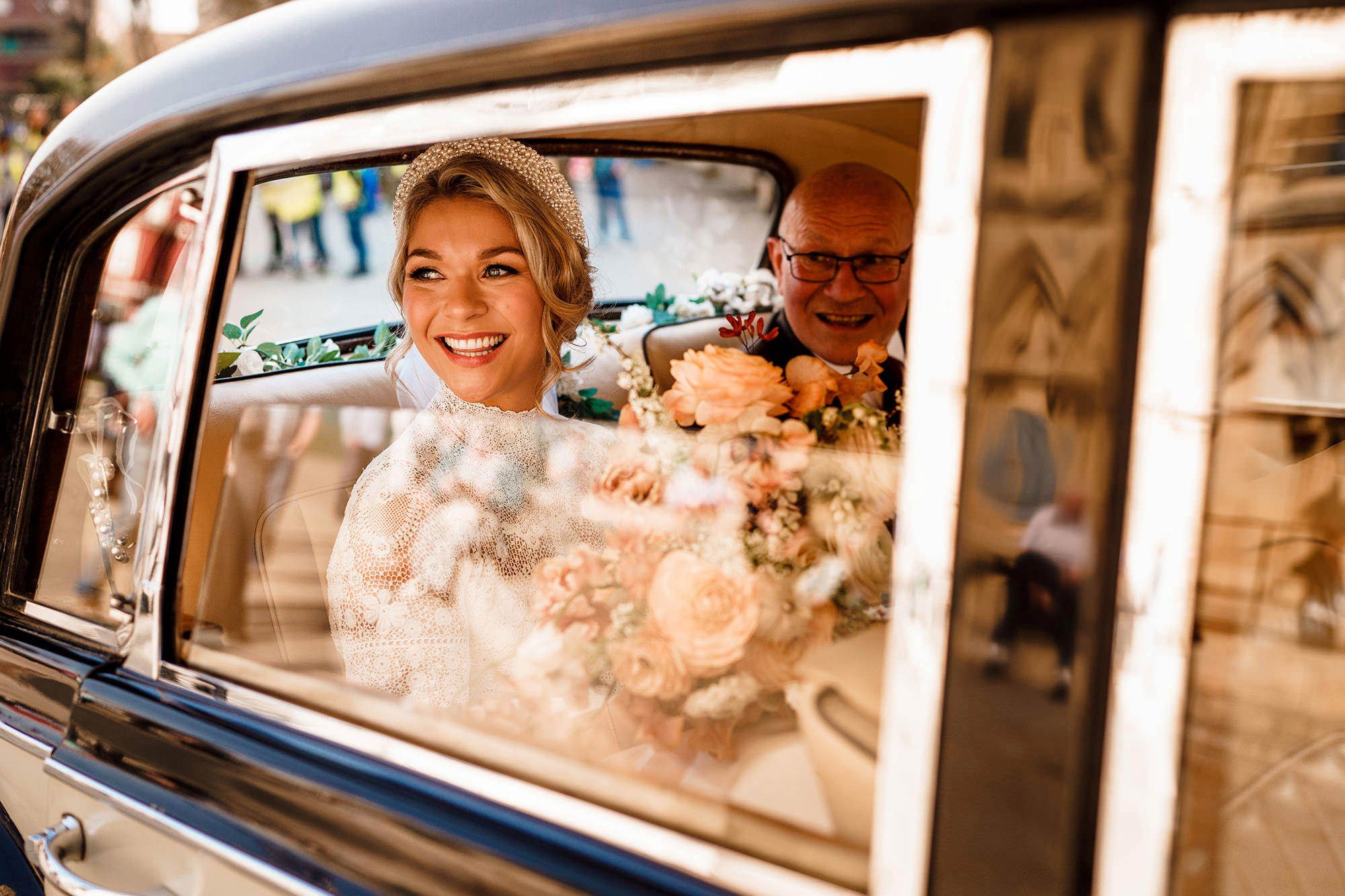Captured through the window of a vintage wedding car, a bride smiles and her father is just visible over the top of the bouquet she's holding. By Stephen Walker Photography