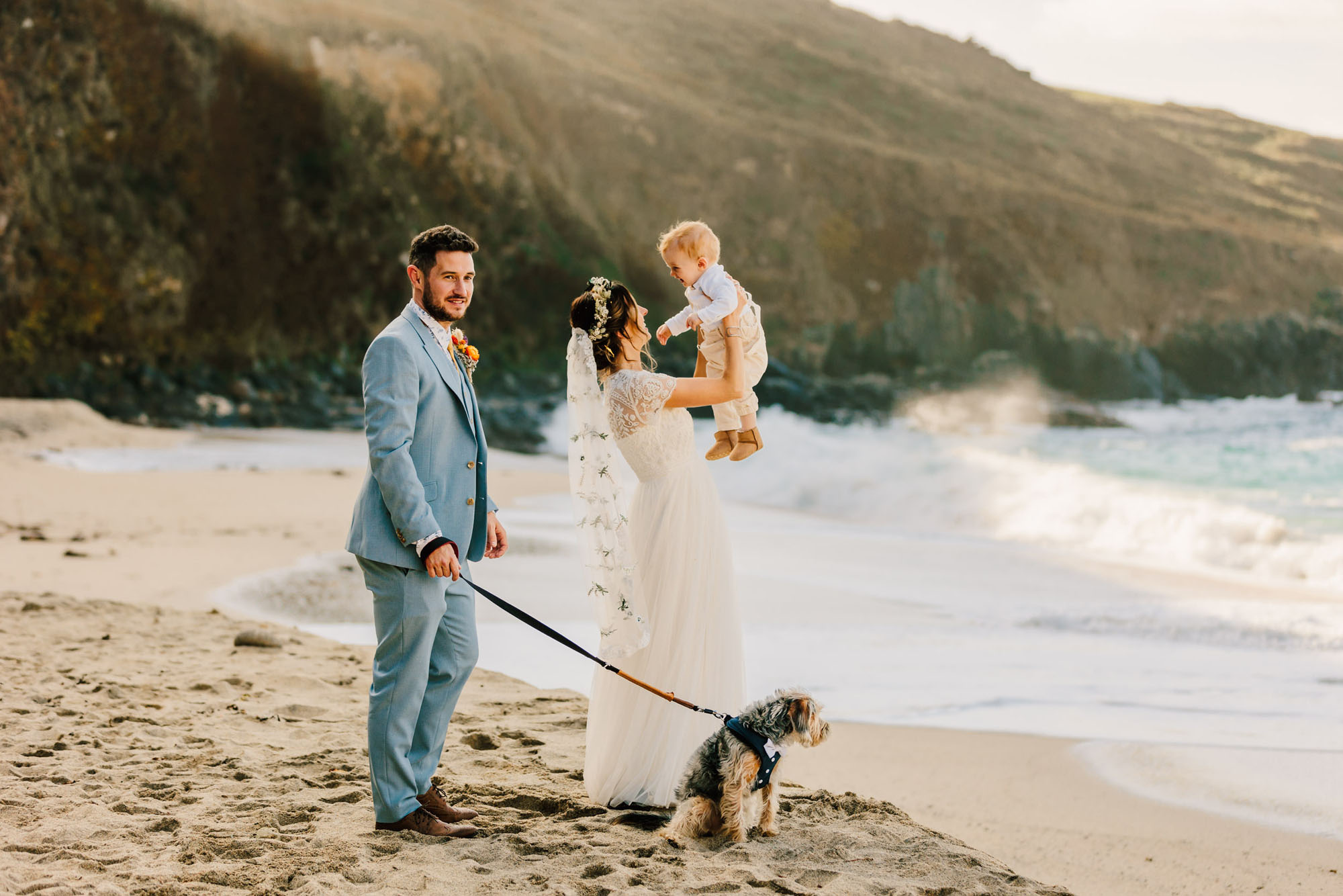 Beautiful wedding day family photo on a beach. The groom holds their dog on a lead, and the bride lifts their toddler into the air. They're on a sandy beach with cliffs behind. By Chris Armstrong Photography in Cornwall