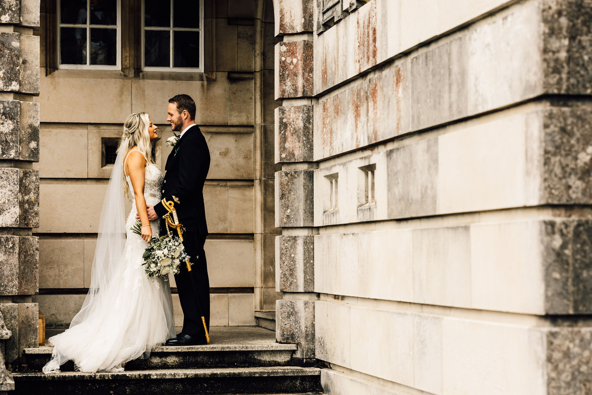 A bride and groom embrace on the steps of a classic wedding venue. He's in full military dress and she's in a white dress. By Chris Armstrong Photography in Cornwall