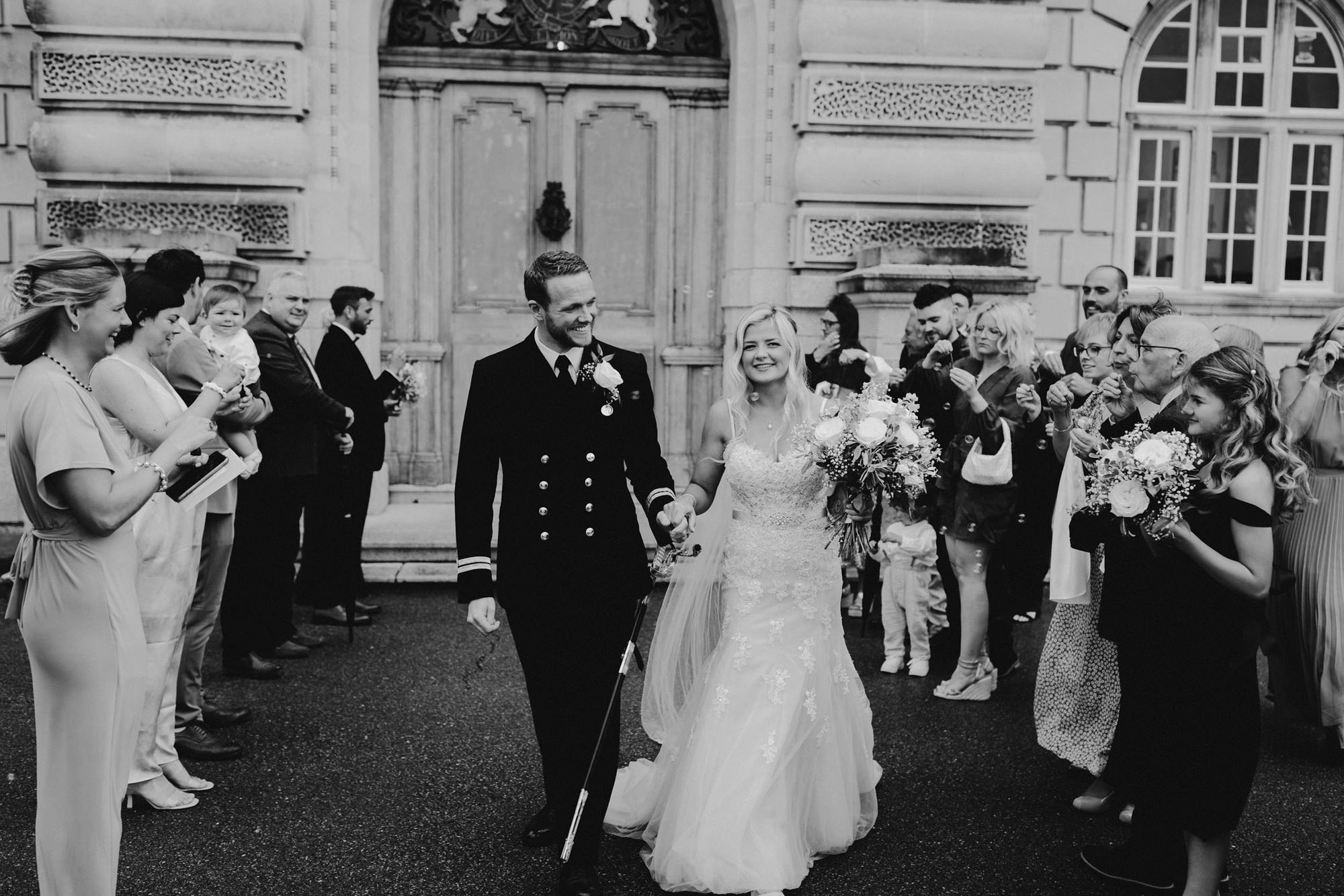 Elegant black and white wedding photograph of a groom and bride as guests blow bubbles instead of confetti. By Chris Armstrong Photography in Cornwall