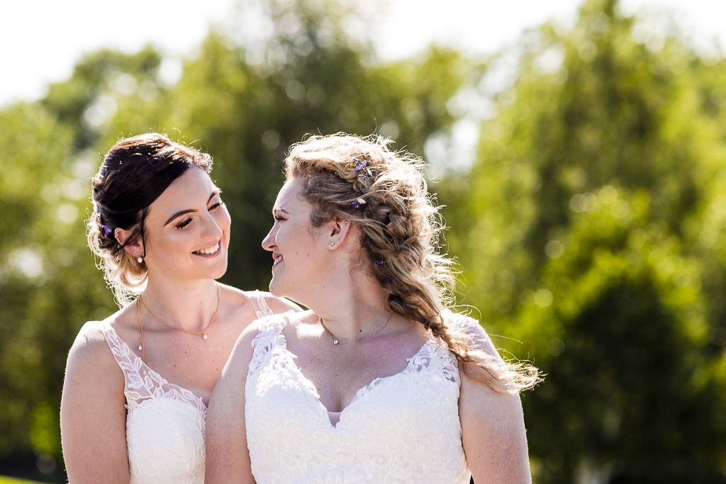 Two brides smile and look into each other's eyes on their wedding day. They're outside in the sunshine and there are trees in the background. By Ross Willsher Photography