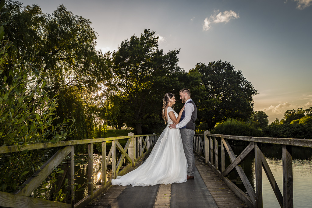 Newlyweds stand on a wooden bridge as the sun begins to set behind them. The bride on the left is wearing a wedding dress with a long skirt which is spread out on the bridge behind her. Ross Willsher Photography