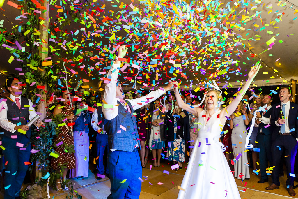 Super colourful confetti shot. A groom and bride carry on dancing as confetti cannons go off and they're showered with colourful paper squares! By Ross Willsher Photography in Essex