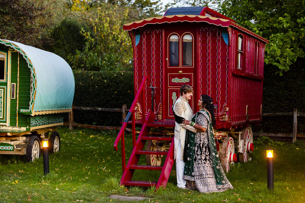 Newlyweds in Indian dress stand by a gypsy caravan holding hands. By Ross Willsher Photography in Essex