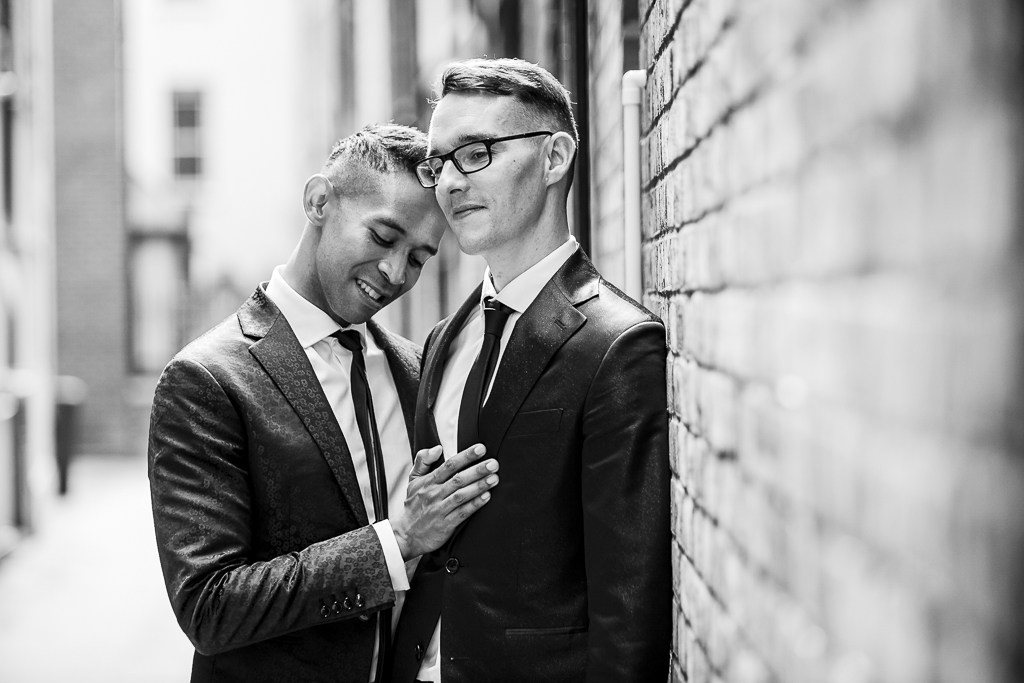 Black and white photography of an LGBTQ couple. They're wearing dark suits and slim ties, and the person on the left has their hand on their partner's chest. By Ross Willsher Photography