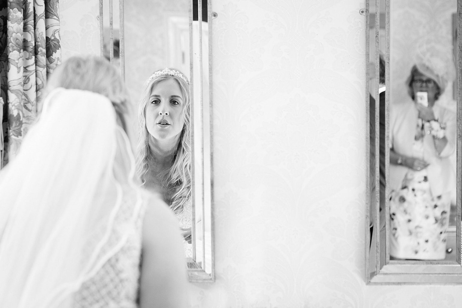 A bride checks her makeup in the mirror while her mum is caught taking a photo on her phone through the other mirror. By 166 Photography