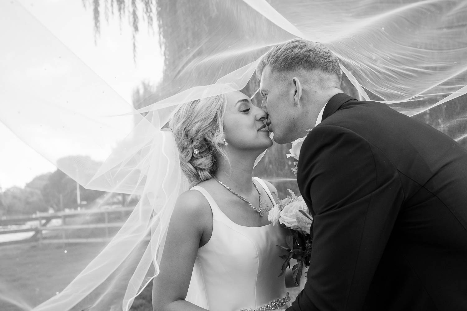 Black and white image of a bride and groom kissing underneath a white veil. With 166 Photography
