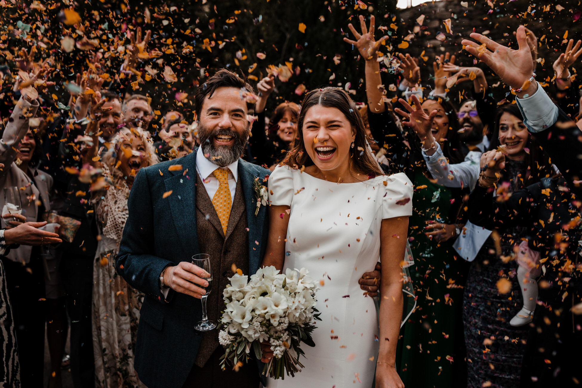 A groom and bride smile as guests throw petal confetti. By Tom Hodgson Photography