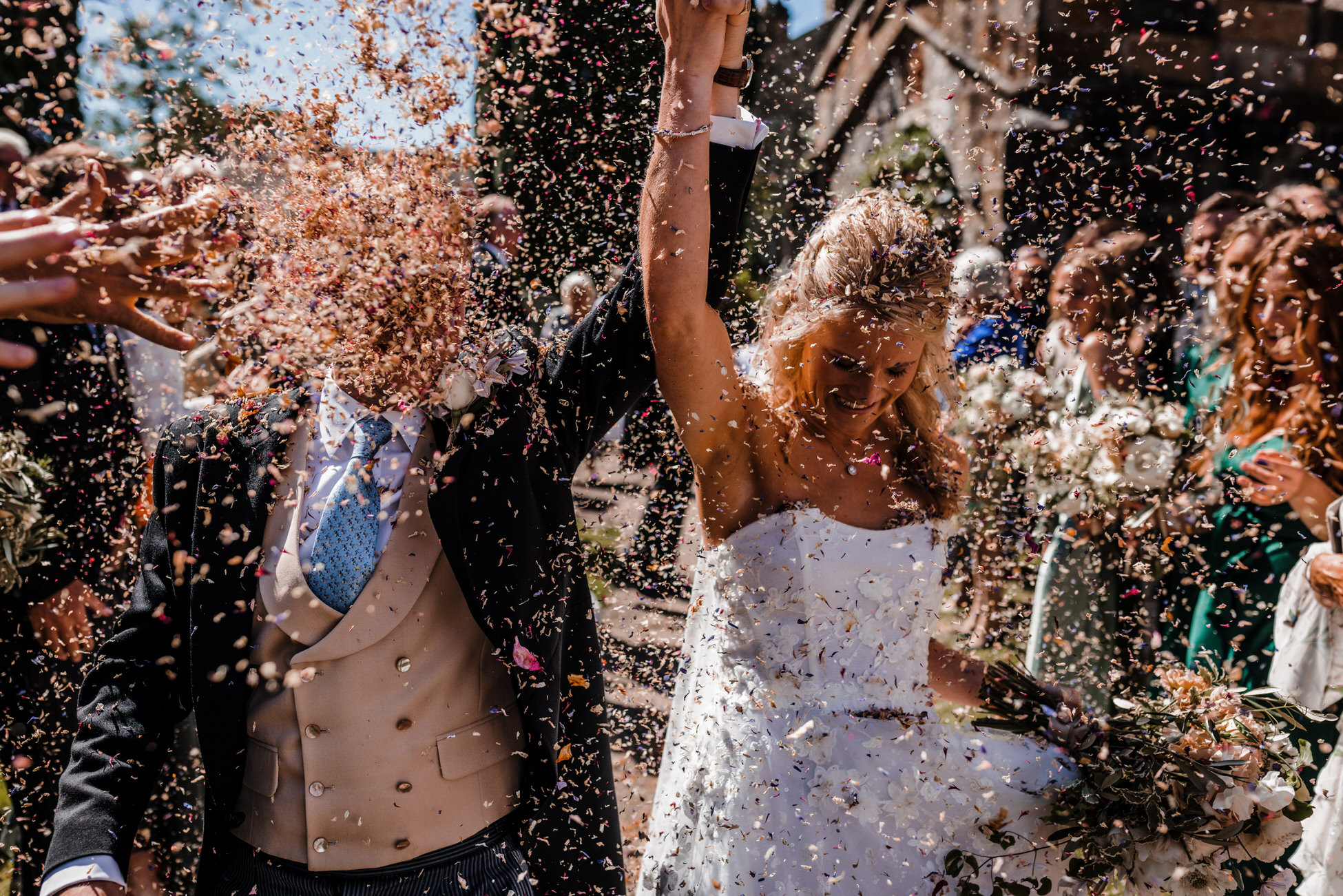 Brilliant confetti photo. Groom Tom gets a face full of dried petal confetti as he holds Anna's hand in the air. Tom Hodgson Photography