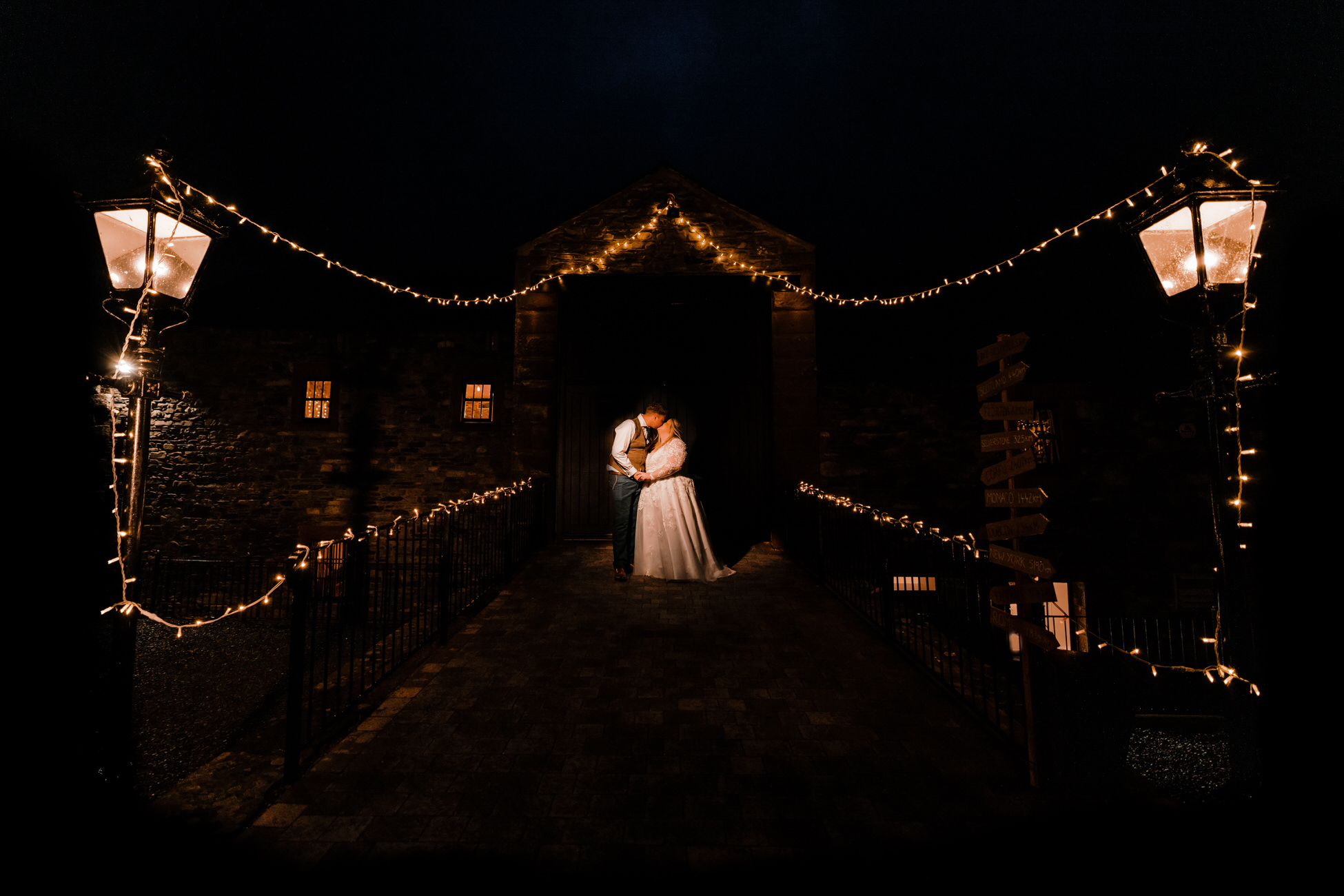 Dramatic dark wedding photo of Leanne and Callum. They hold each other under a canopy of festoon lights. By Tom Hodgson Photography in the Peak District