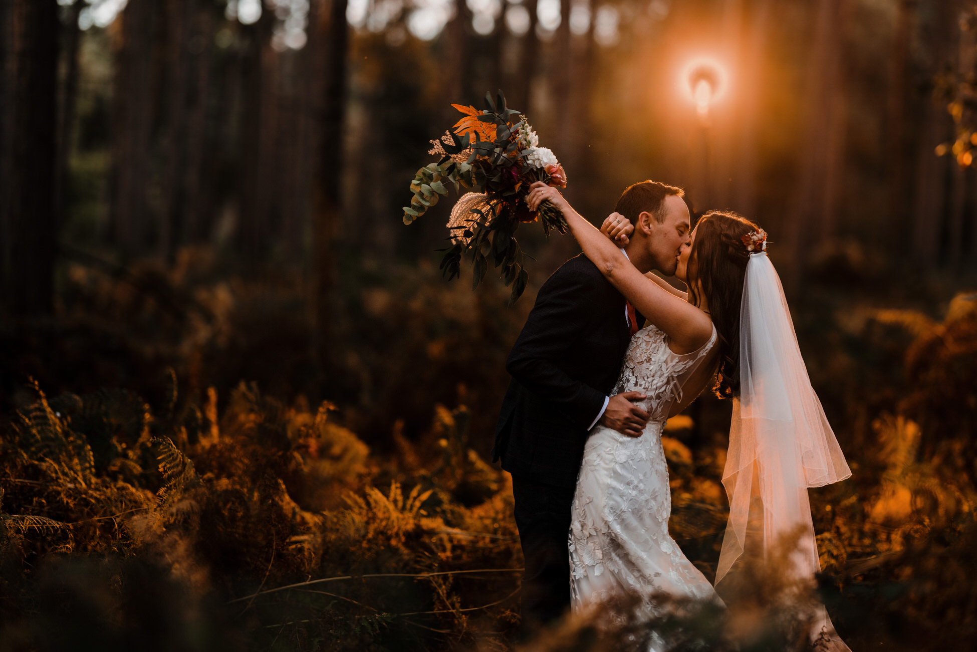 Glorious golden hour wedding photography! Emma and Sam share a hug and a kiss as the sun sets. There's a woodland behind them and she's holding her bouquet with her arms around Sam's shoulders. By Tom Hodgson Photography