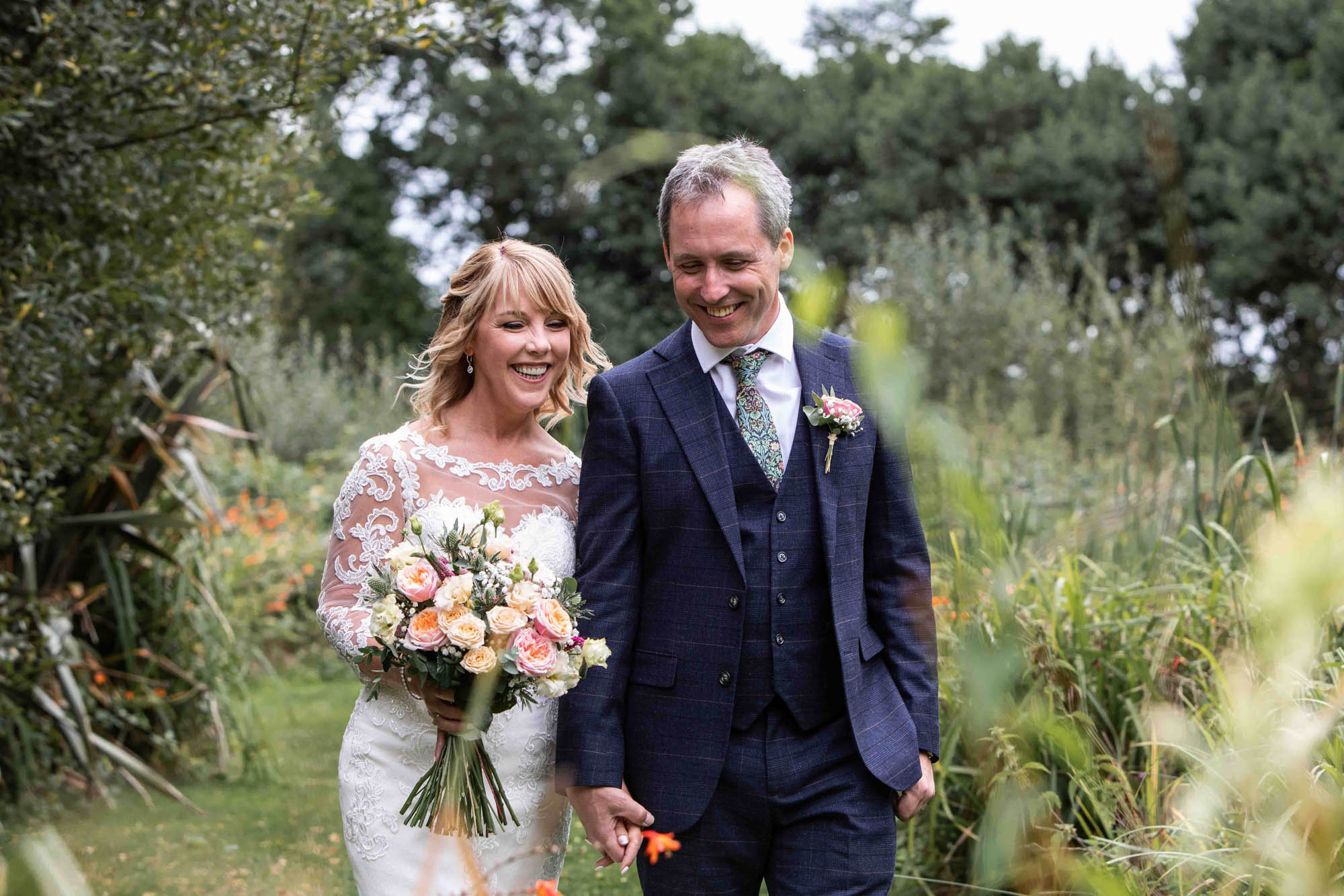 Mature couple on their wedding day. The bride is carrying a bouquet of pale roses and wearing a lace embroidered dress. The groom's in a dark blue suit with waistcoat and they're holding hands. By Snapdragon Photography