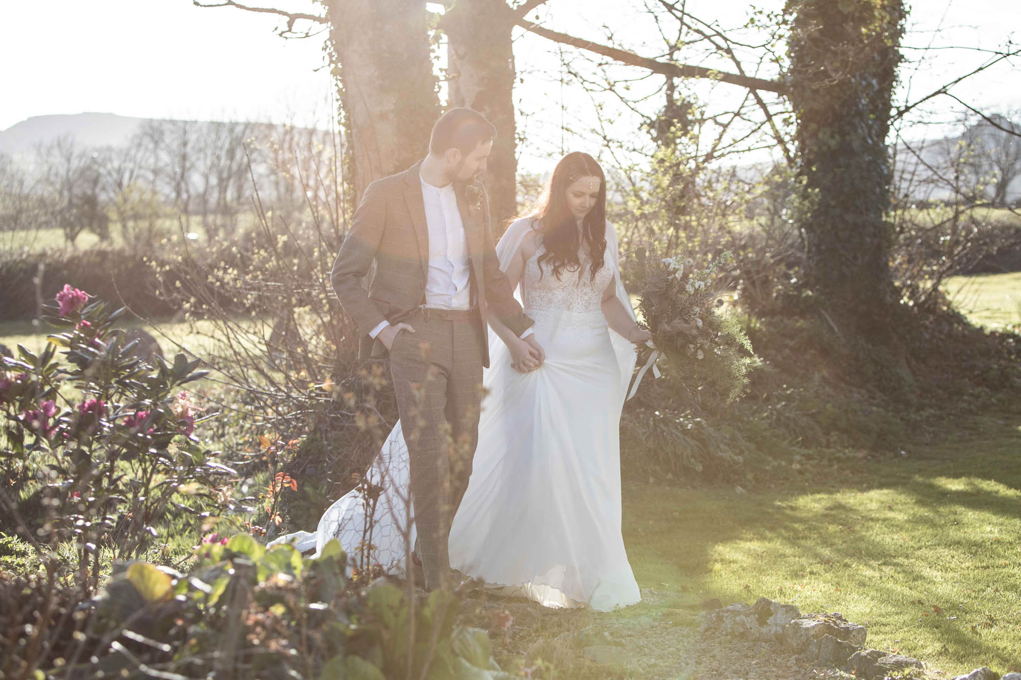 Hazy bright sunlit photograph of a bride and groom walking in a woodland. By Snapdragon Photography