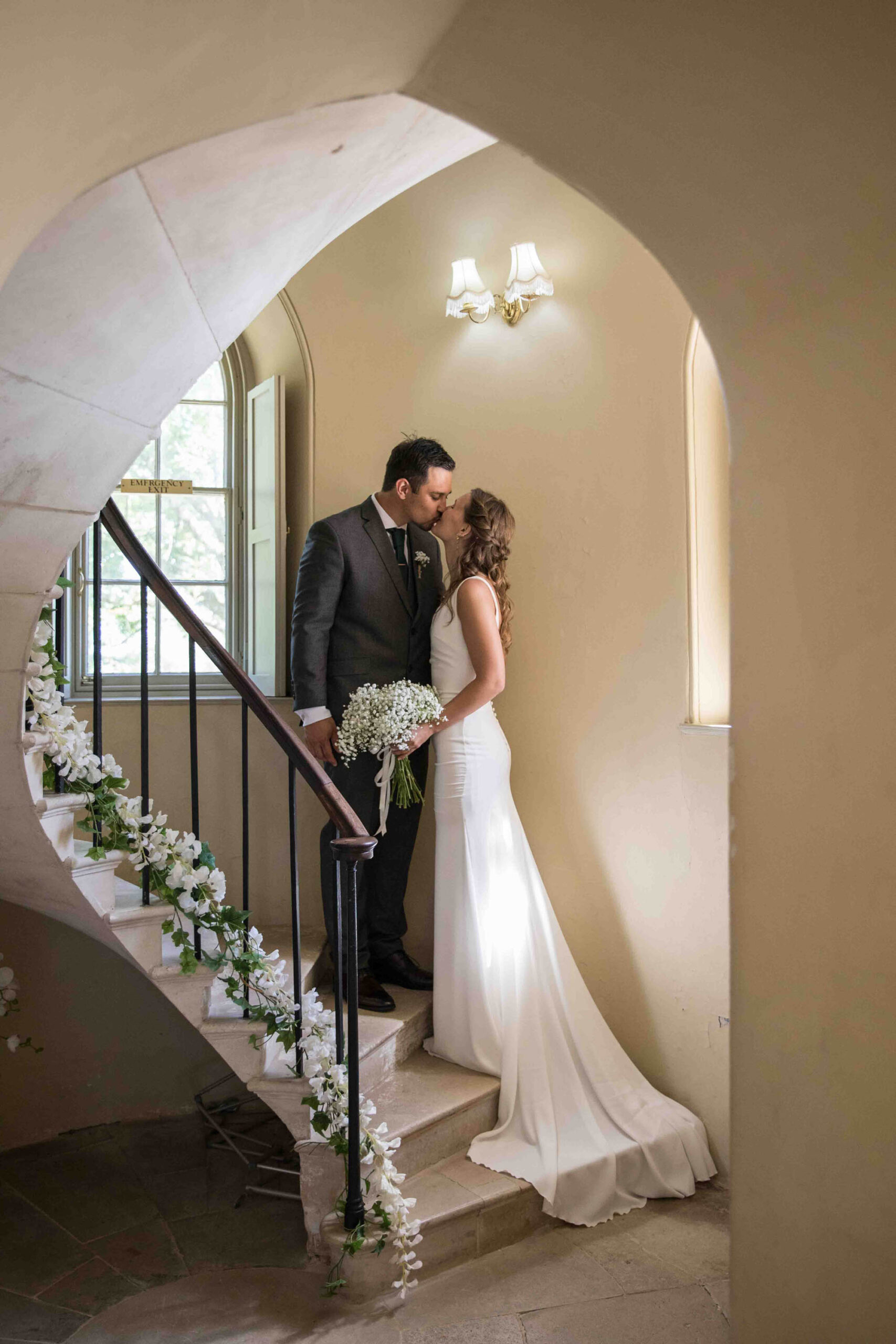 Elegant wedding portrait taken from an archway in a stately home. There are flowers lining the staircase where a bride and groom are sharing a kiss. Snapdragon Photography is based in Devon