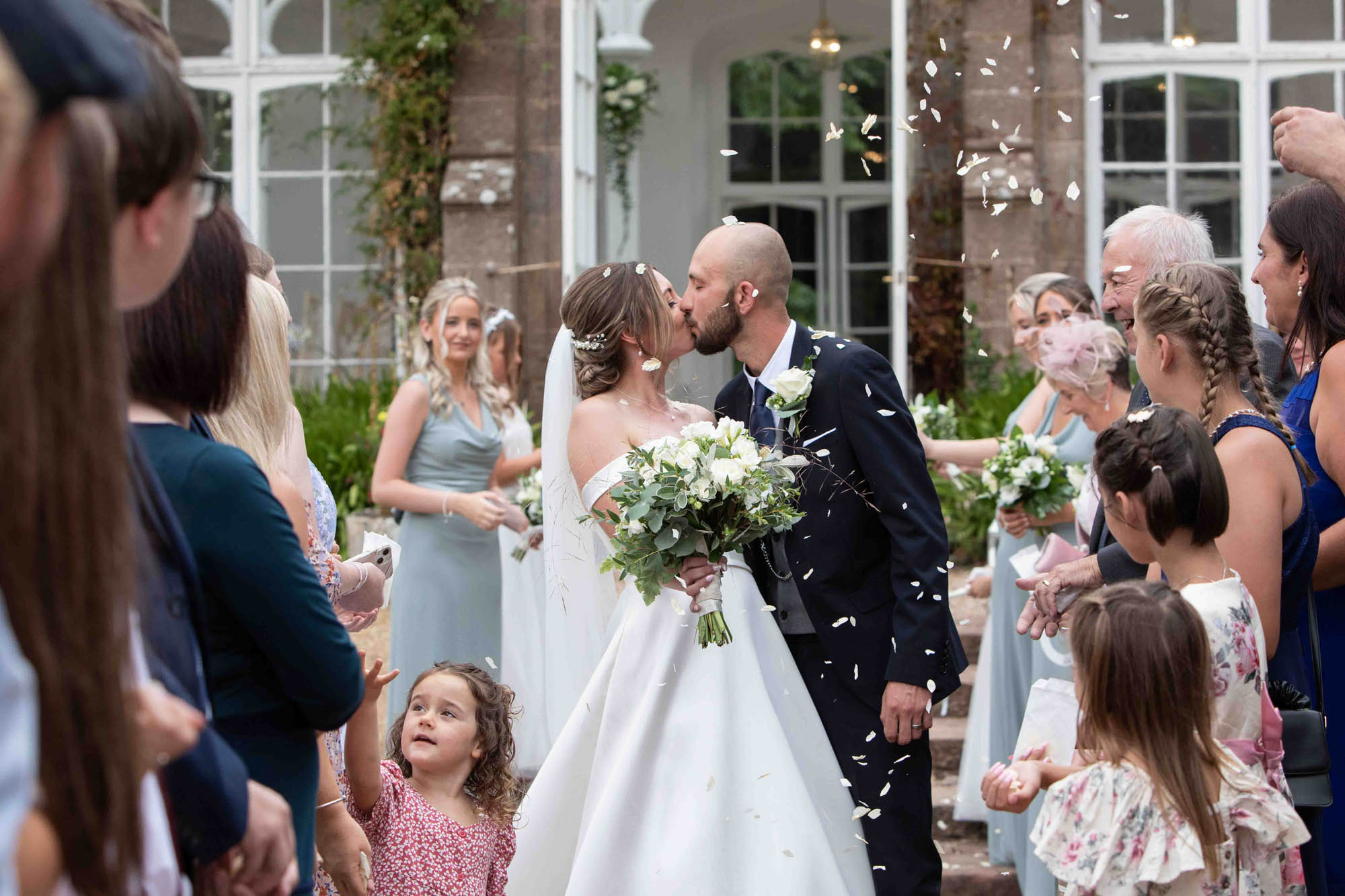 A bride and groom kiss on their wedding day as guests throw confetti. Snapdragon Photography Devon
