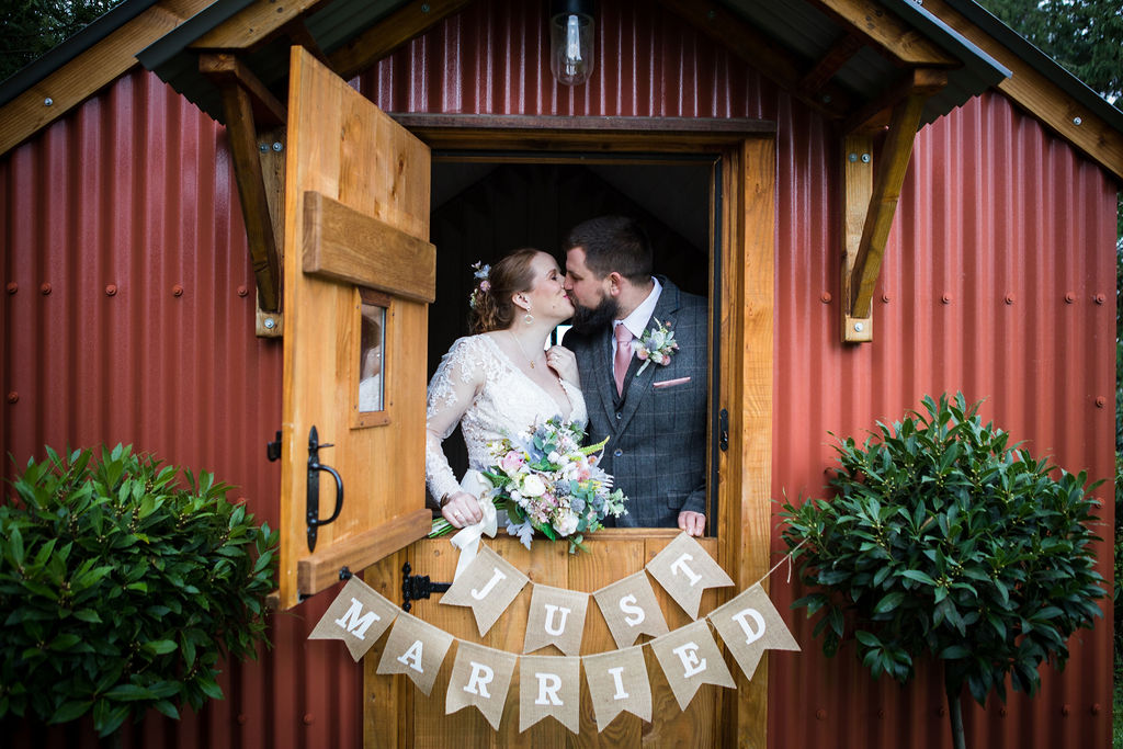 Newlyweds kissing by a stable door strung with Just Married bunting at Little Silver Weddings