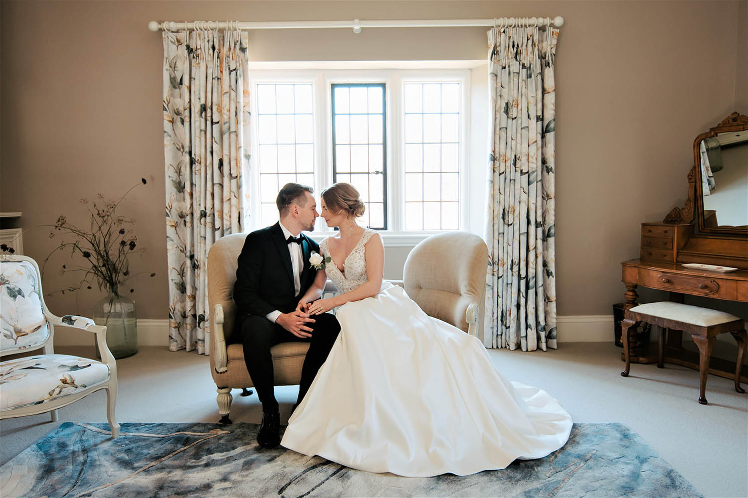 Romantic wedding image of a bride and groom sat on an elegant chair before a bright window.