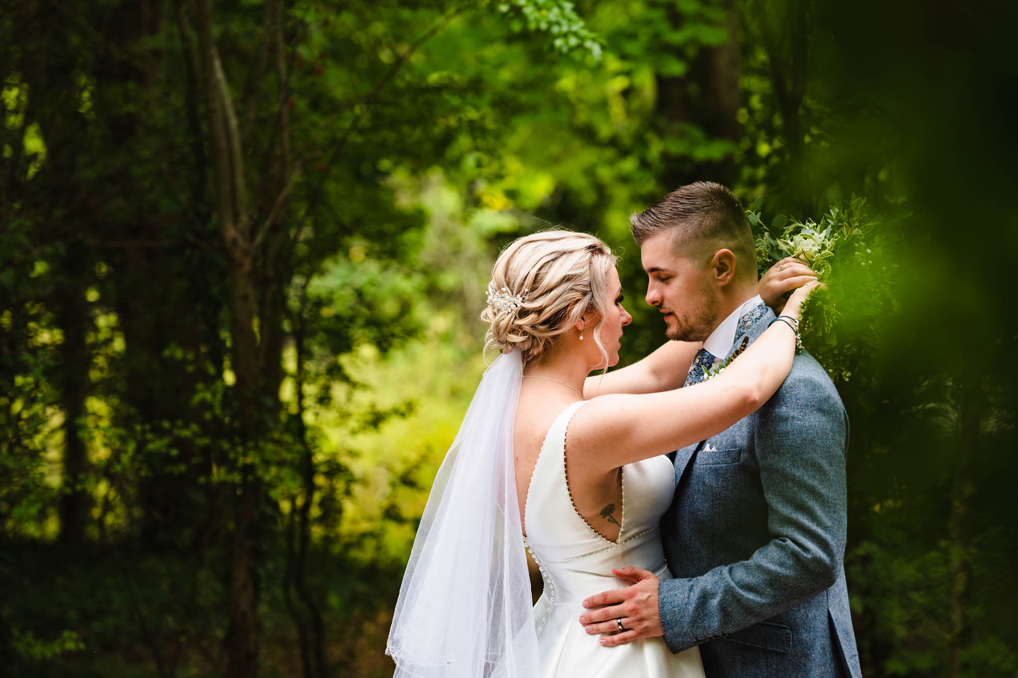 Romantic picture of newlyweds in woodland. The bride has her arms around the groom and he's holding her waist