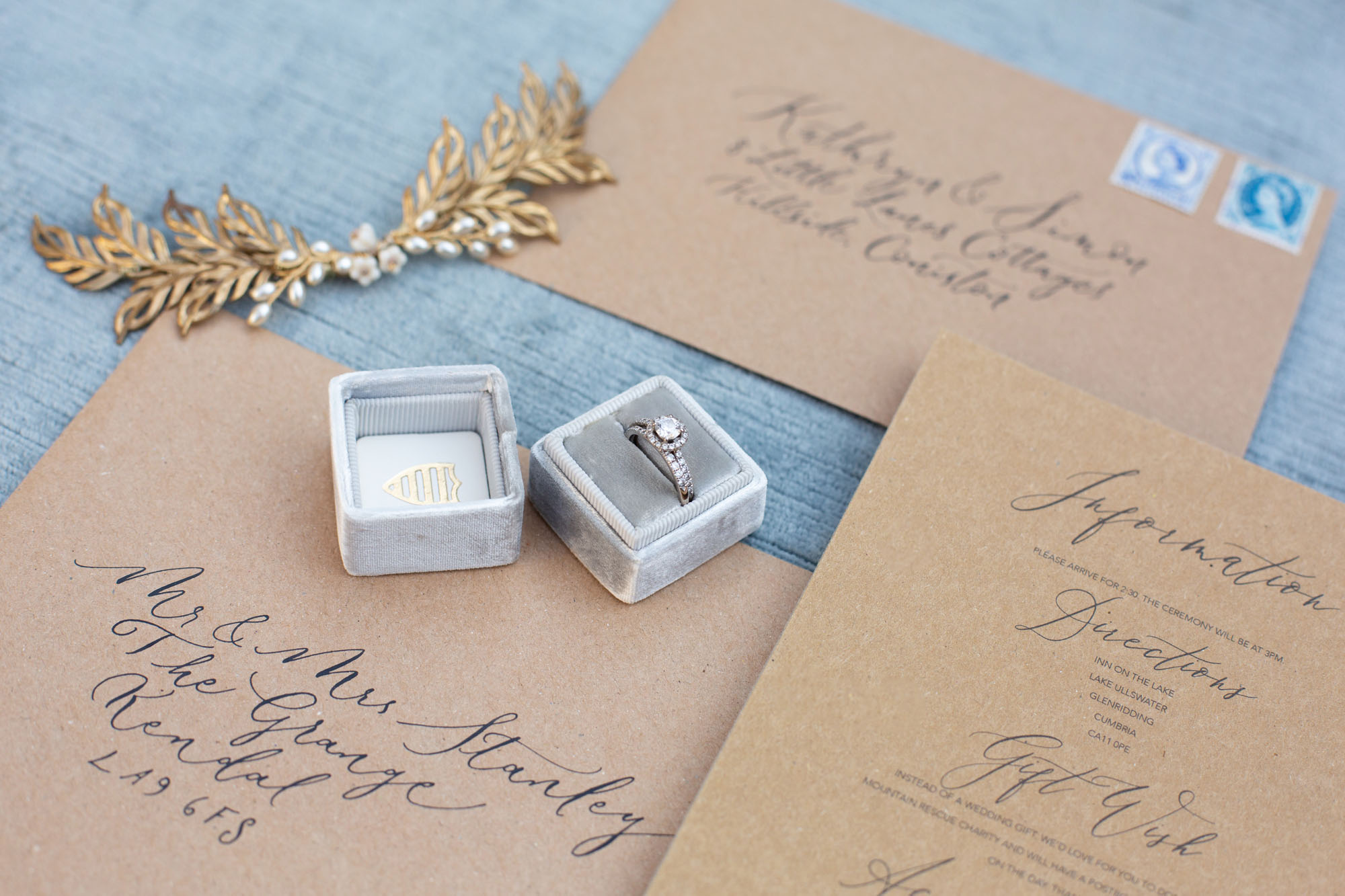 An open wedding ring box with a halo diamond ring sits on a calligraphy envelope with a gold hair accessory to one side. By Jessica Reeve Photography