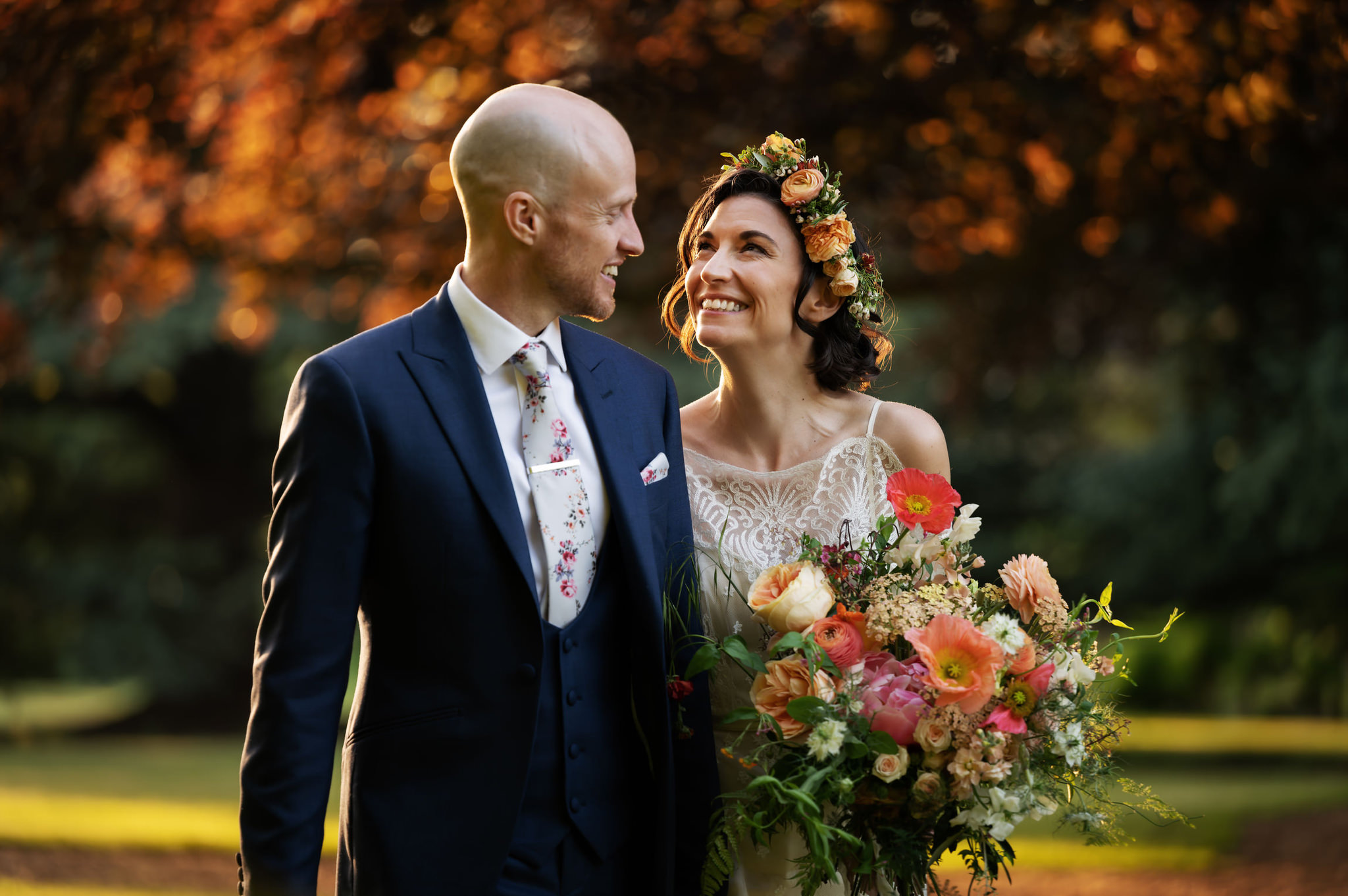 Buckinghamshire wedding photographer Andy Sidders. A couple look into each others eyes. She's holding an autumnal bouquet. He's wearing a smart suit. The light is golden and the background is a garden in autumn.