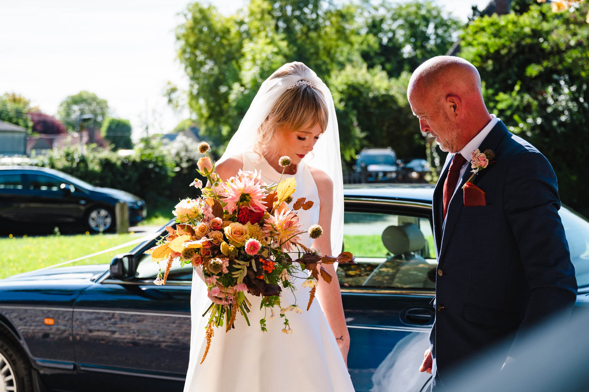 A bride stands by her wedding car holding an autumnal bouquet in front of her. 