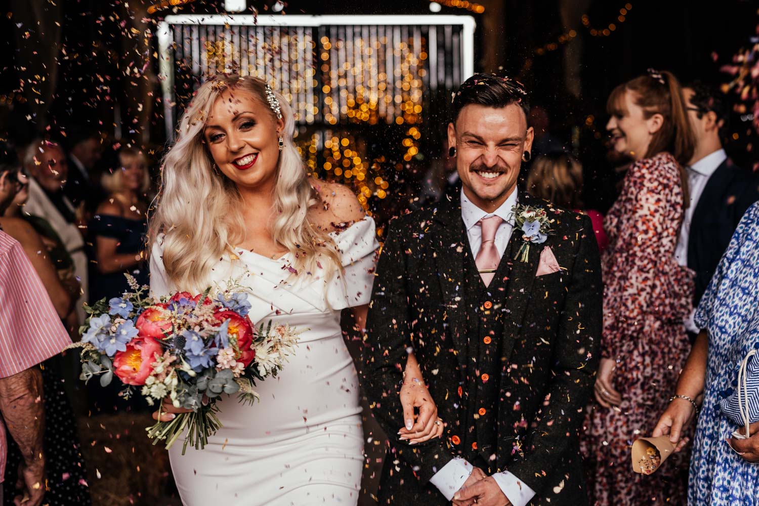 Newlyweds smiling as confetti falls in front of them. The bride is holding a colourful bouquet of flowers. Joss Denham Photography