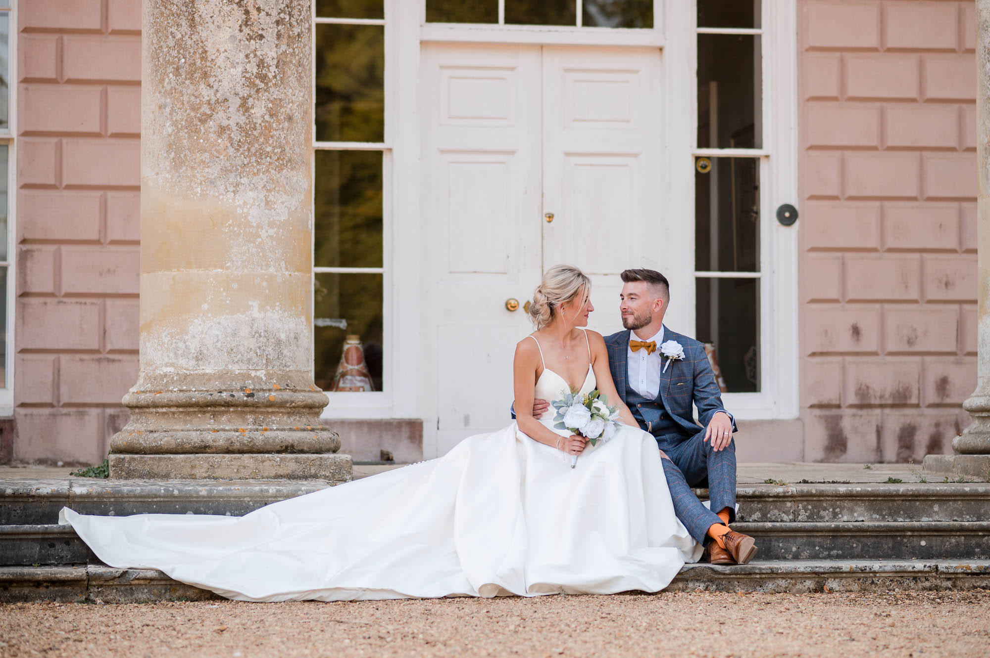 Bride and groom sat on the steps of Hale Park wedding venue. She's wearing a dress with a full train and holding flowers. He's in a tan bowtie with orange socks. By Libra Photographic, Dorset