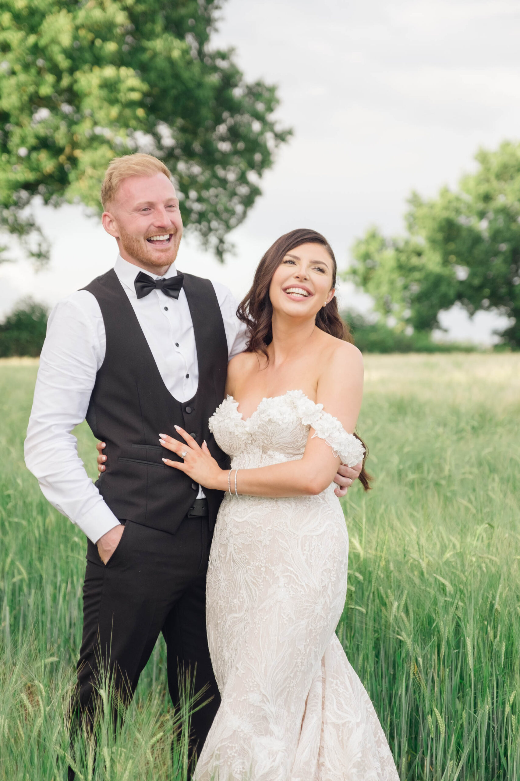 Groom and bride with a green cornfield behind them. He's in black bowtie and waistcoat with a white shirt. She wears a Bardot neck wedding dress. By Jordan Fox Photography