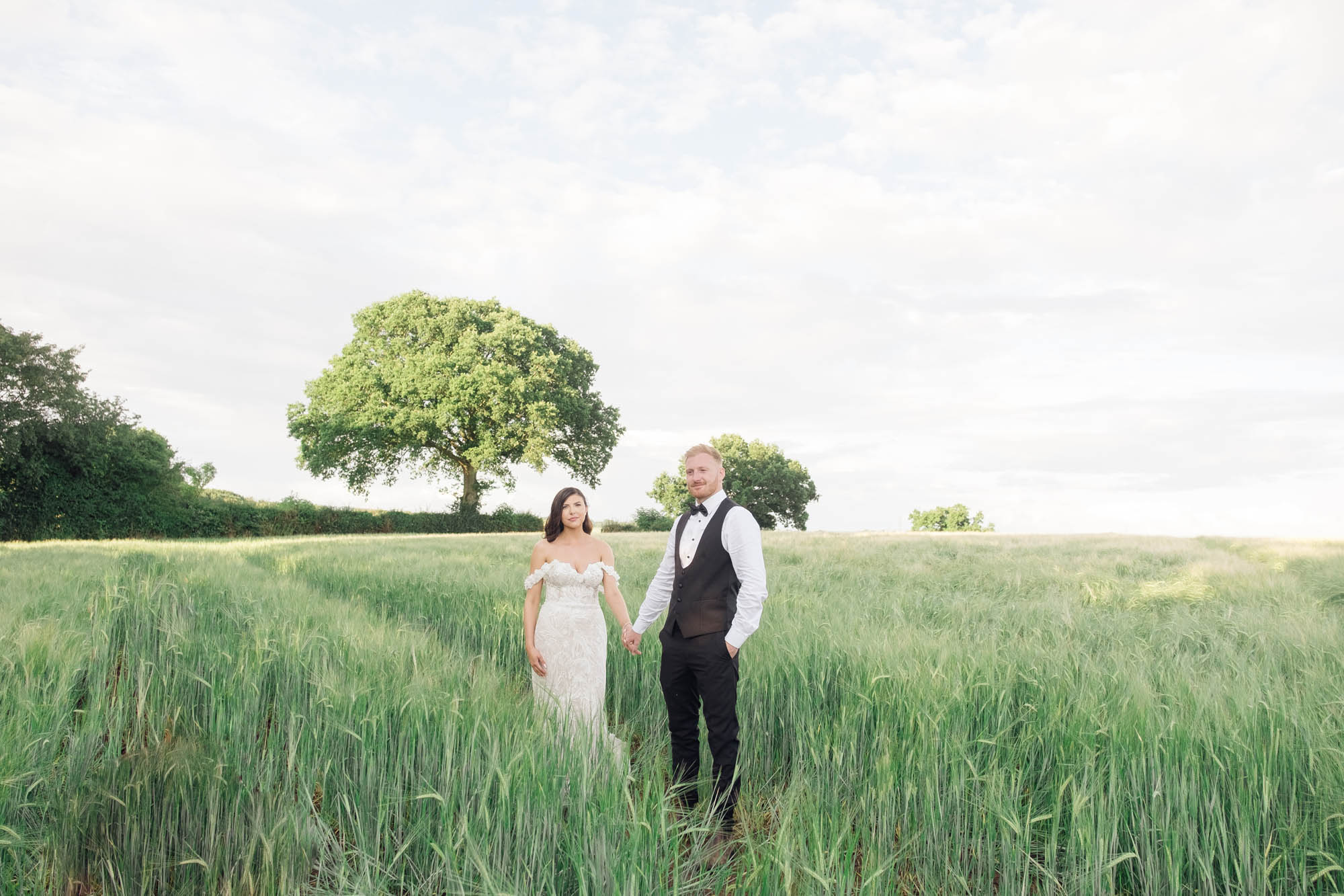 Wide angle wedding photo of a bride and groom in a green cornfield with trees behind them. By Jordan Fox Photography
