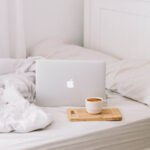 a coffee and a MacBook on a white bed with the duvet rumpled up