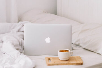 a coffee and a MacBook on a white bed with the duvet rumpled up