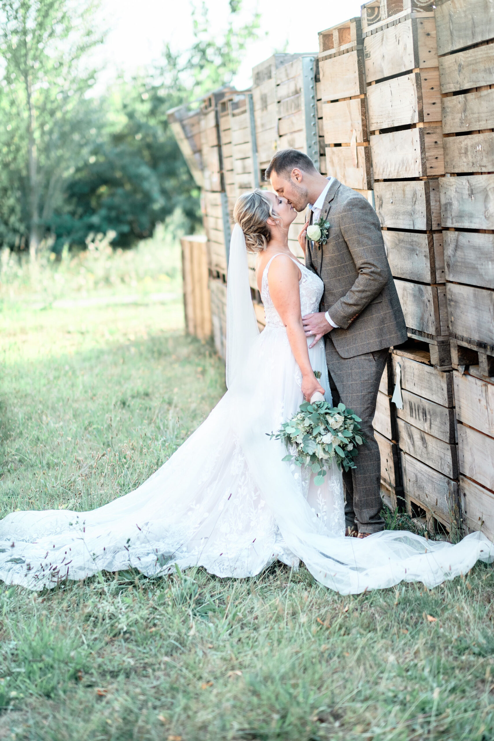 A groom and bride kiss by a rustic wooden fence. He's in a brown suit and she's in a white dress with veil holding a bouquet. By Howling Basset Photography