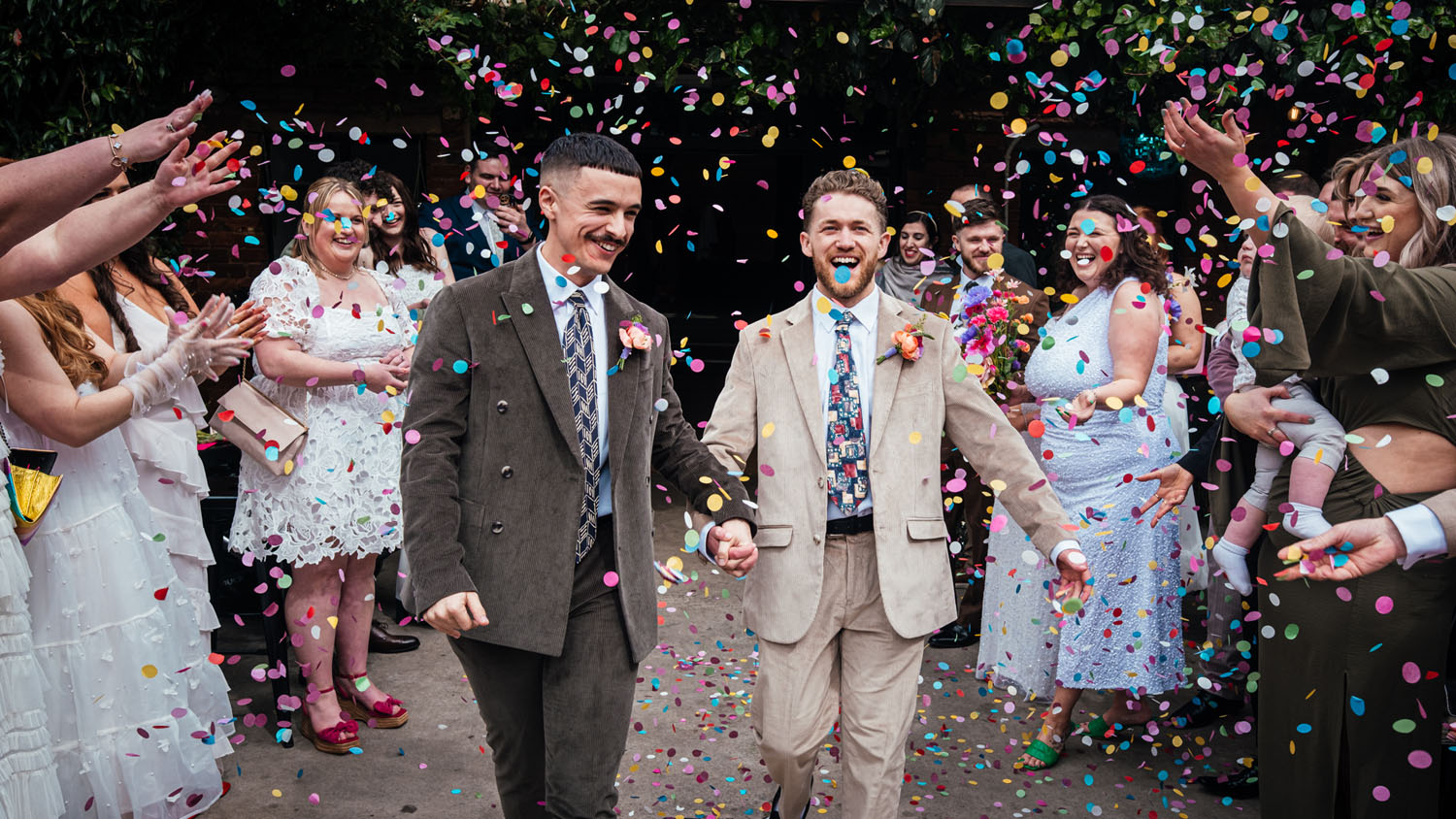Two grooms in a burst of colourful confetti by documentary wedding photographer Simon Dewey