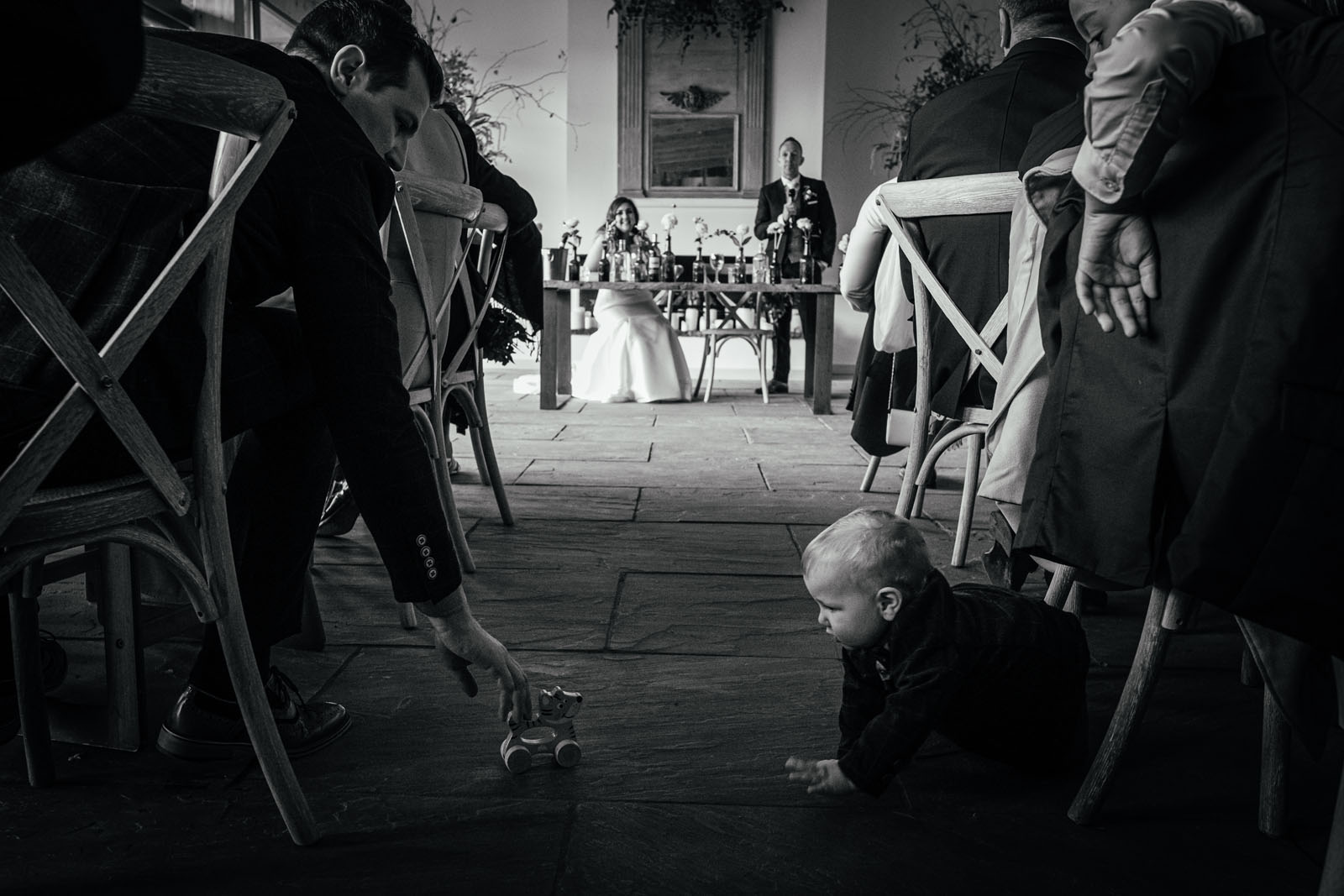 A toddler plays with a wooden toy in the aisle of a wedding venue while the bride and groom watch, smiling, at the end. Black and white documentary wedding image by Simon Dewey Photography in Derby UK