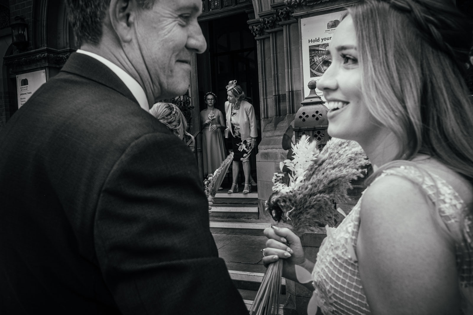 A man and woman stand outside a wedding venue, talking and smiling in the foreground. The woman's holding a bouquet of dried grasses and florals. By documentary photographer Simon Dewey