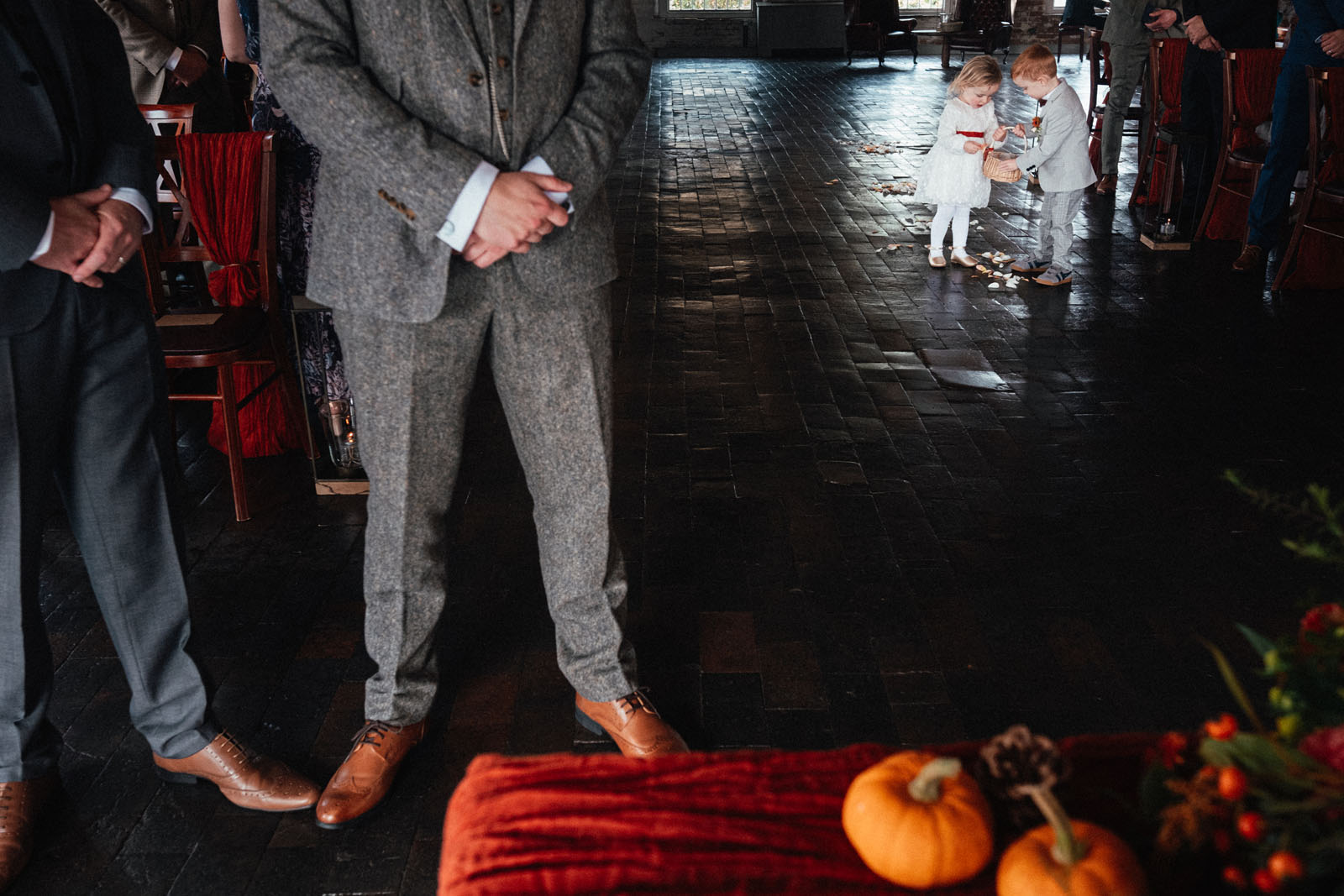 A layered documentary wedding photograph showing a dressed table with rust coloured cloth and mini pumpkins in the foreground. The groom stands with clasped hands, in a grey suit with tan shoes. In the background a tiny flower girl and page boy huddle over a basket of petals to scatter along the aisle. By Derby documentary wedding photographer Simon Dewey