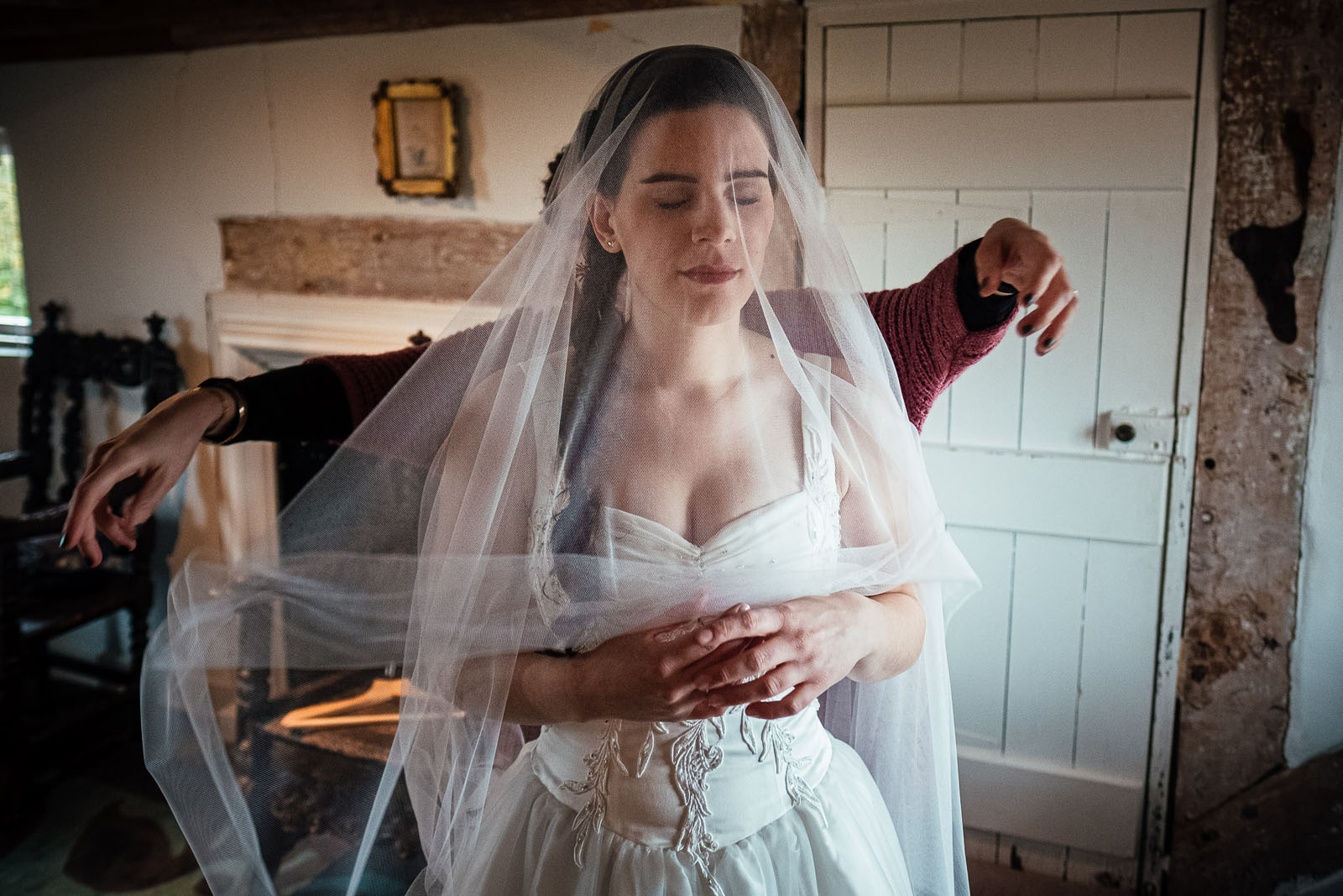 A bride stands with closed eyes and clasped hands as someone behind her drapes the veil over and around her. Documentary UK wedding photography by Simon Dewey