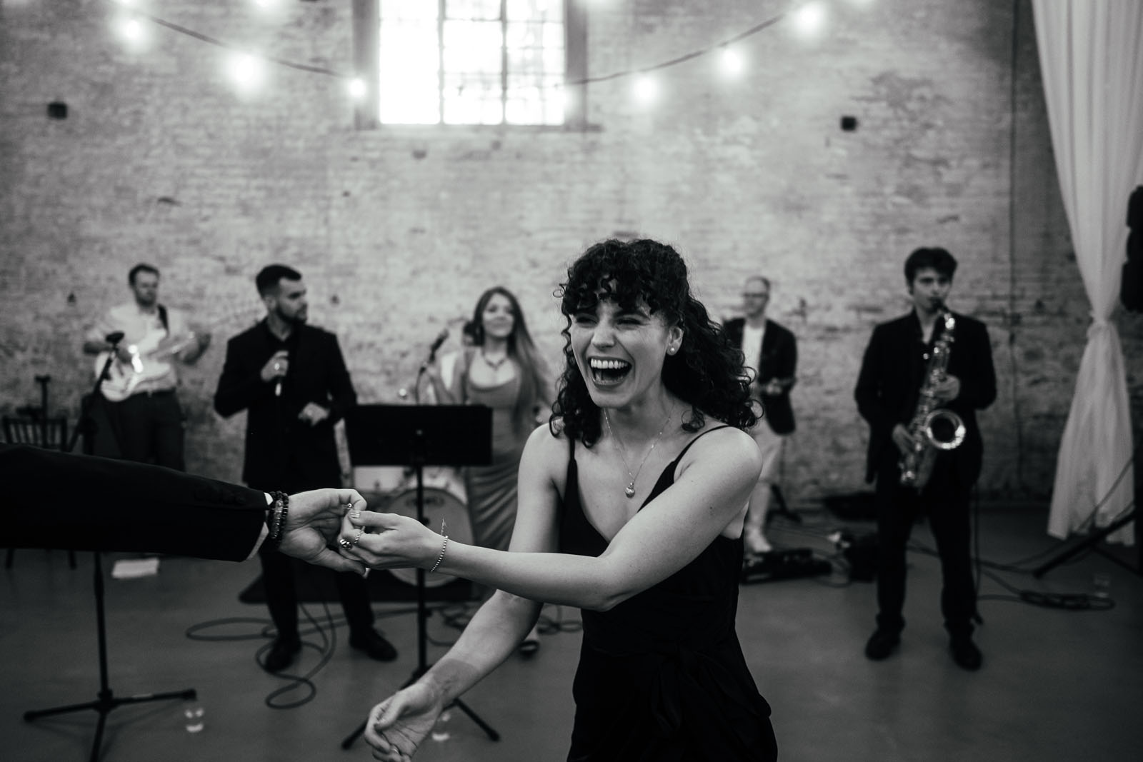 A band plays in the background of a Derby wedding venue while a woman dances, laughing and holding her partner's hand. UK documentary wedding photography by Simon Dewey