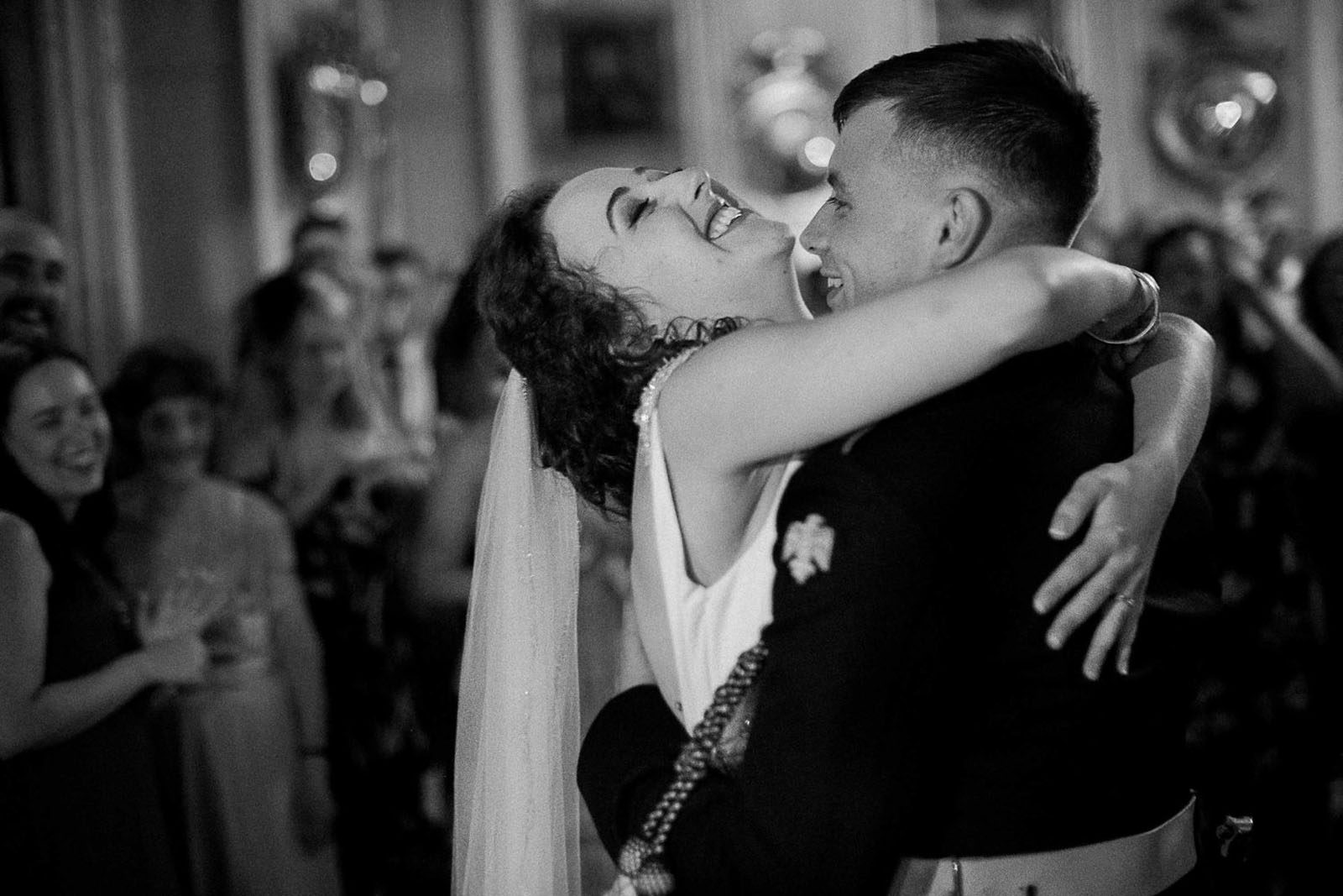 A bride grins and holds her groom close as they dance at a wedding reception. Guests smile in the background. She's wearing a veil and he's in military dress. Simon Dewey Photography