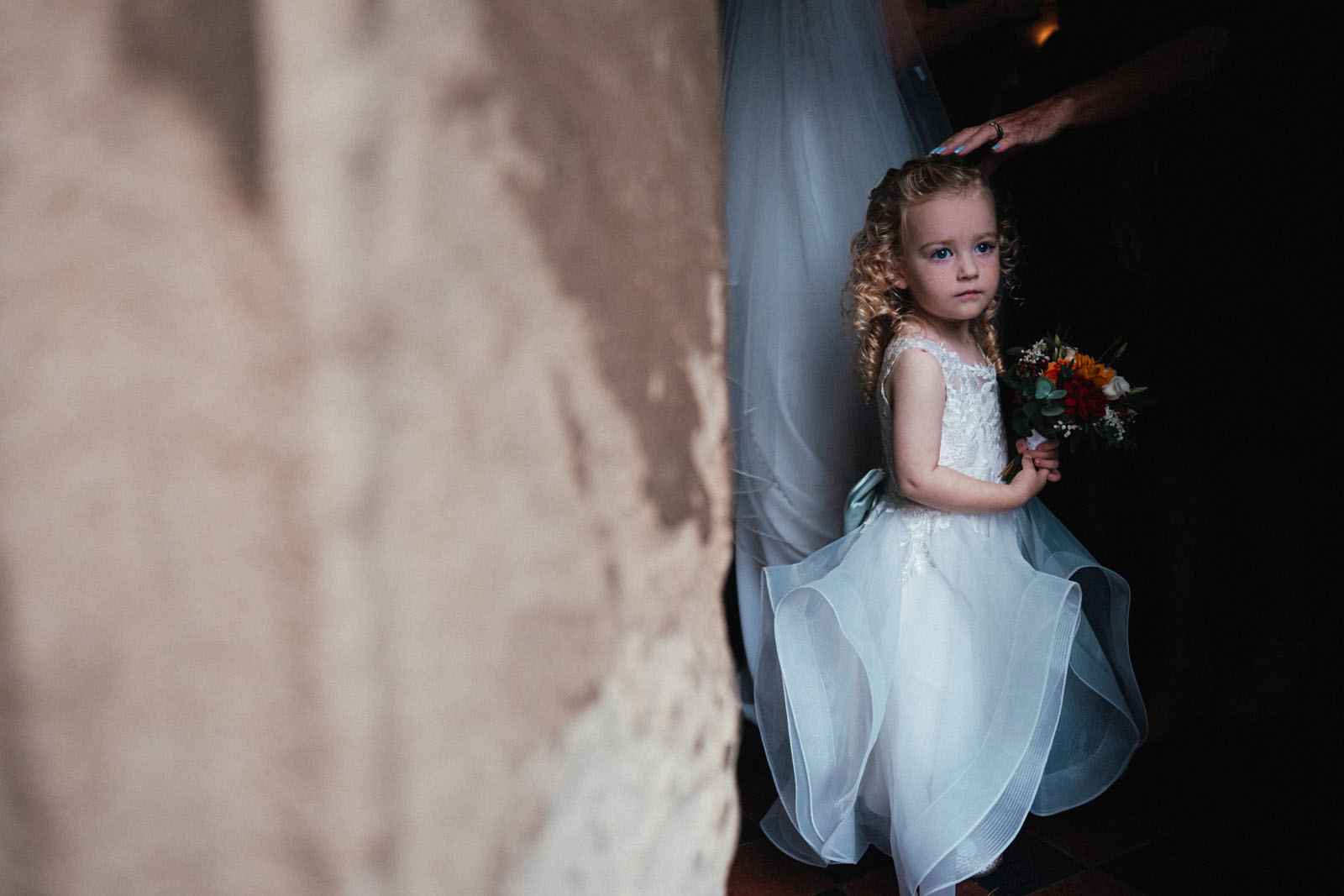 A little bridesmaid looks past the camera as an adult places a hand on her head. She's carrying a bouquet of autumnal flowers. With Simon Dewey, UK documentary wedding photographer