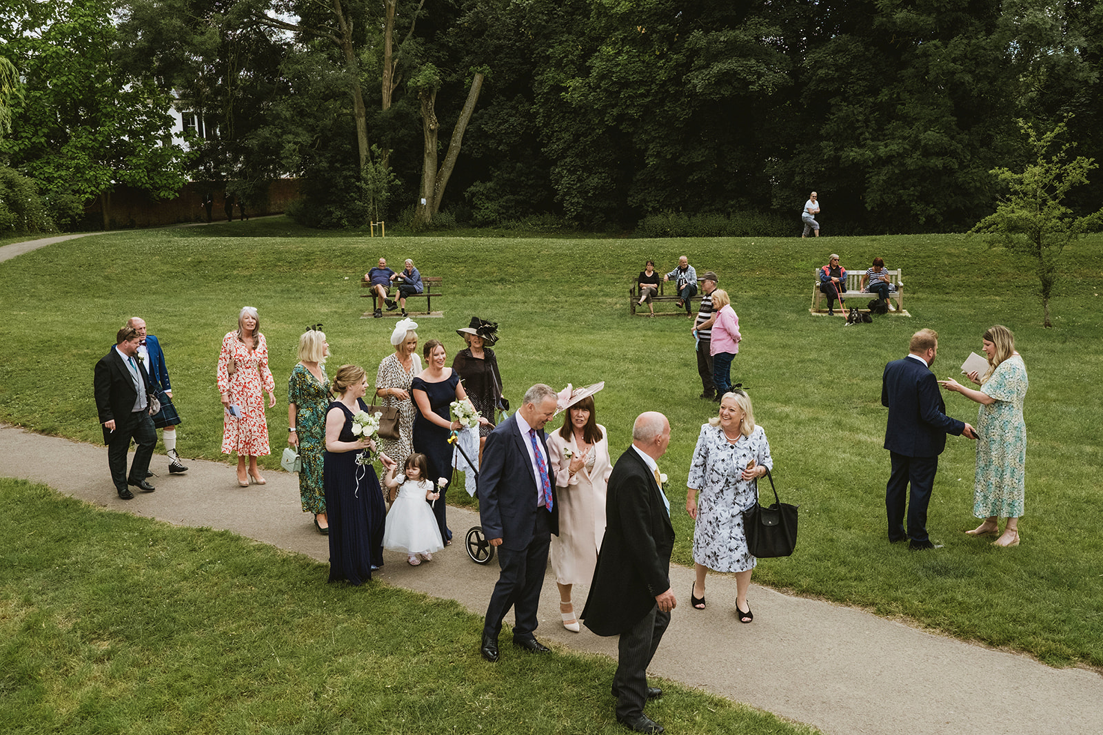 Guests walk along a path through a riverside park dressed in wedding attire while people on benches watch them go by. York Place Studios in Buckinghamshire