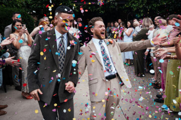One of two grooms at a wedding looking thrilled to spot a guest in the crowd during the confetti throw - Simon Dewey Photography